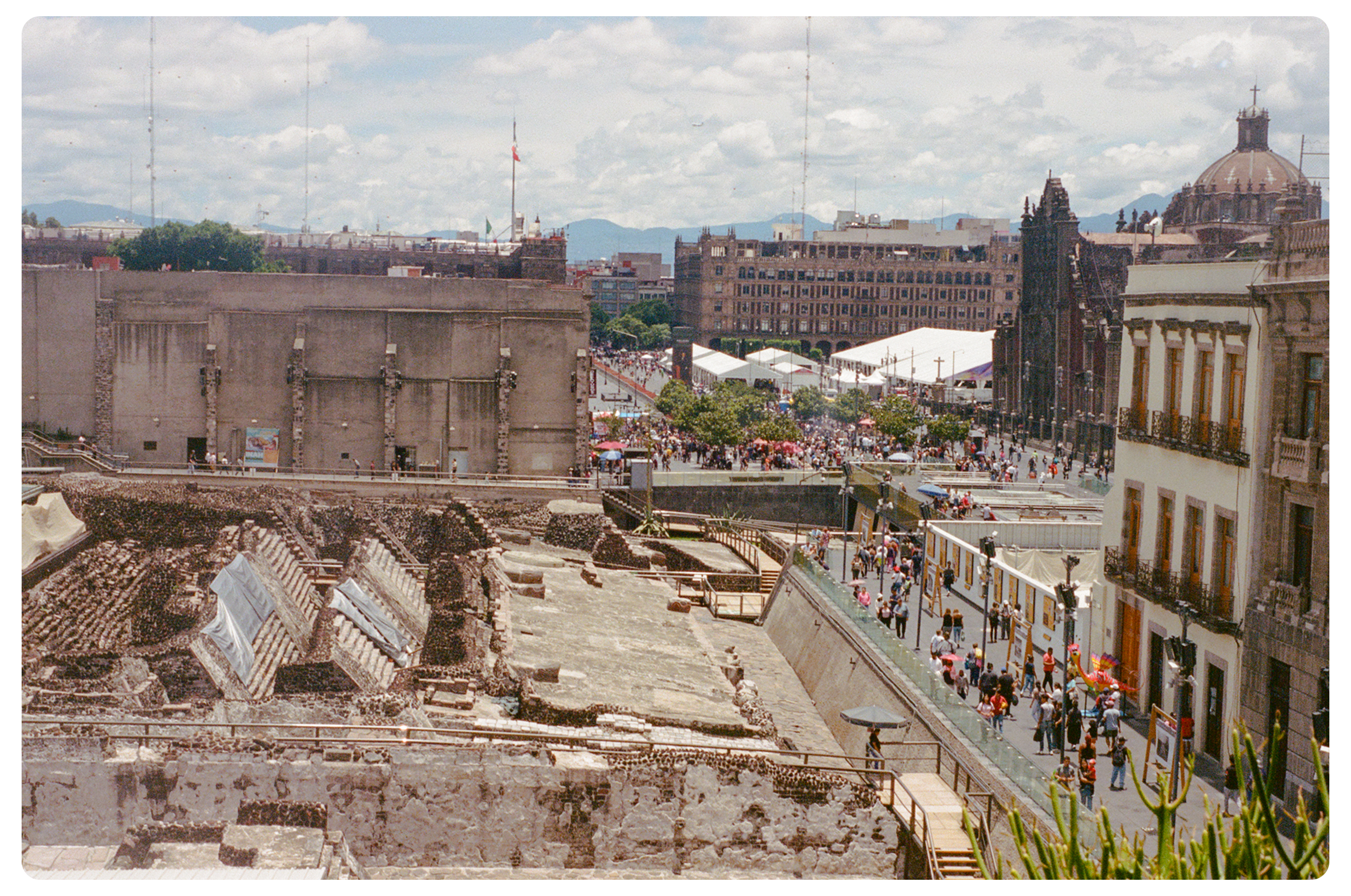 An aerial view of a historic archaeological site with ruins in the foreground and a busy city plaza with crowds, tents, and buildings in the background.