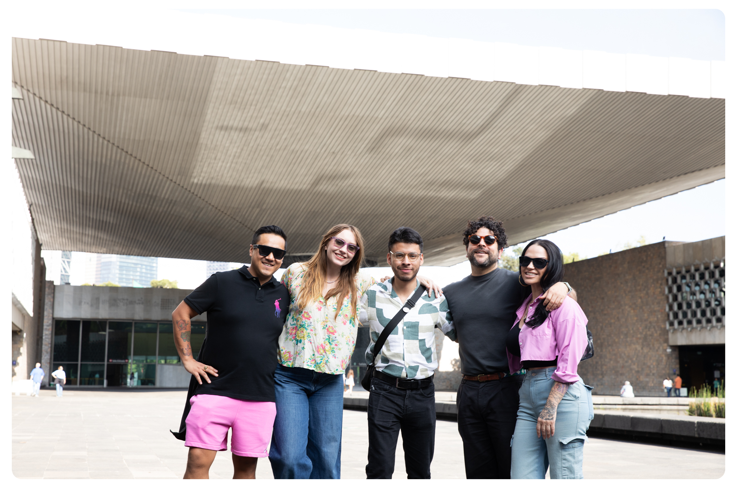 Group of five young adults standing together outdoors in front of modern architecture, smiling, with some wearing sunglasses and casual clothing.
