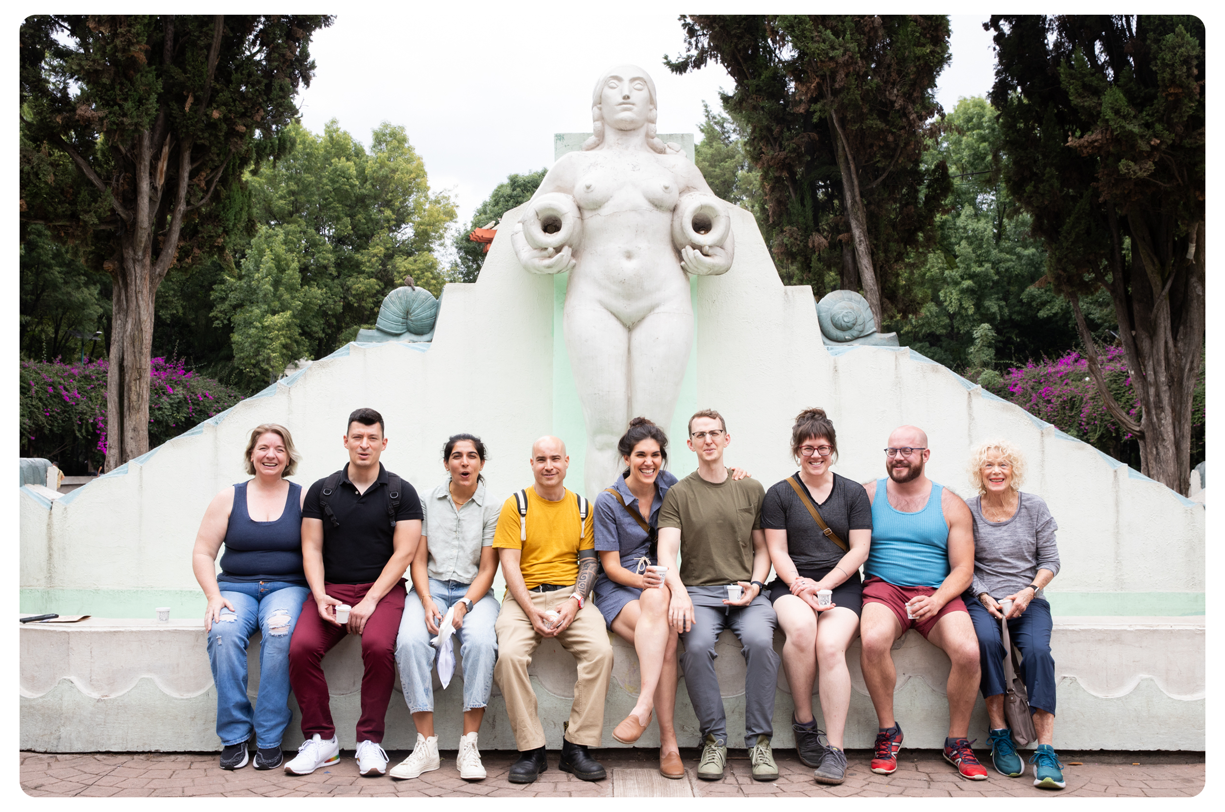 A group of nine people sitting on a concrete ledge in front of a large statue of a woman holding two conch shells. The background features trees and purple flowering bushes.