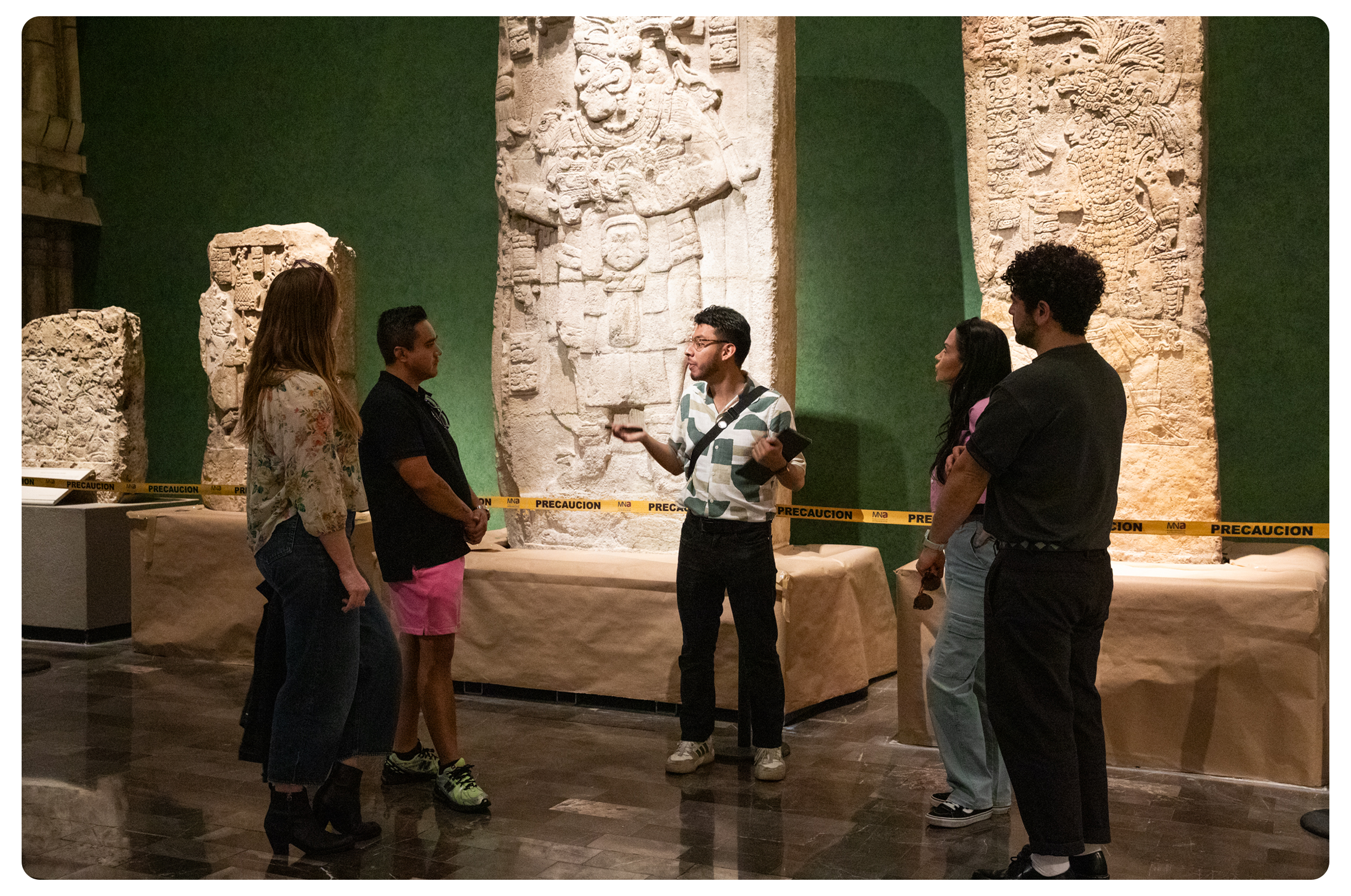 A tour guide explains ancient stone carvings to a group of five visitors inside a museum. The carvings depict intricate figures and patterns, and the setting is marked off with caution tape.