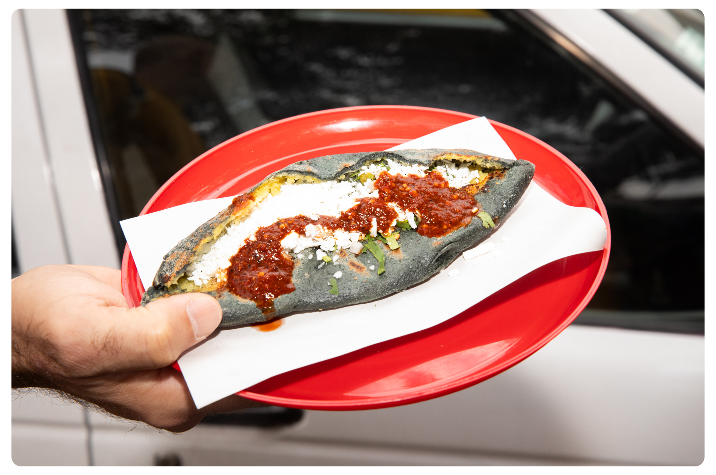 A hand holding a red plate with a black-colored Mexican taco, filled with cheese and topped with red sauce, on white paper, with a blurred car door in the background.