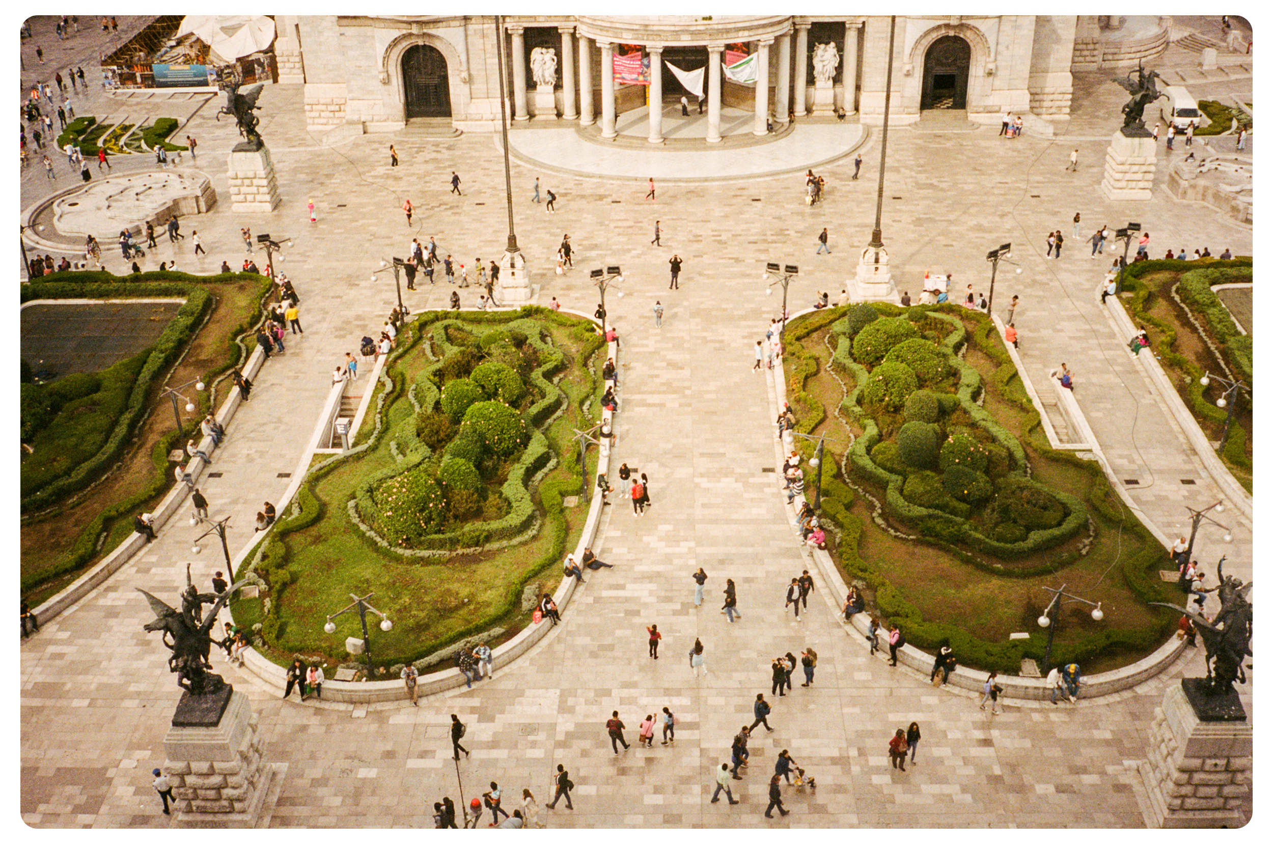 Bird's eye view of a large city square with two ornate, landscaped gardens, foot traffic, statuary, and a grand building with columns in the background.