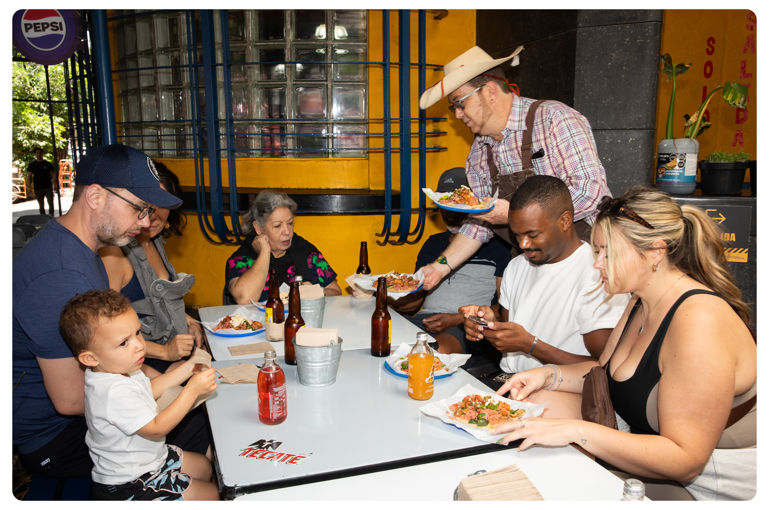 Family gathered at a restaurant table with plates of pizza, soda bottles, and a young boy eating, while a man in a cowboy hat serves food, and others look at their phones or converse.
