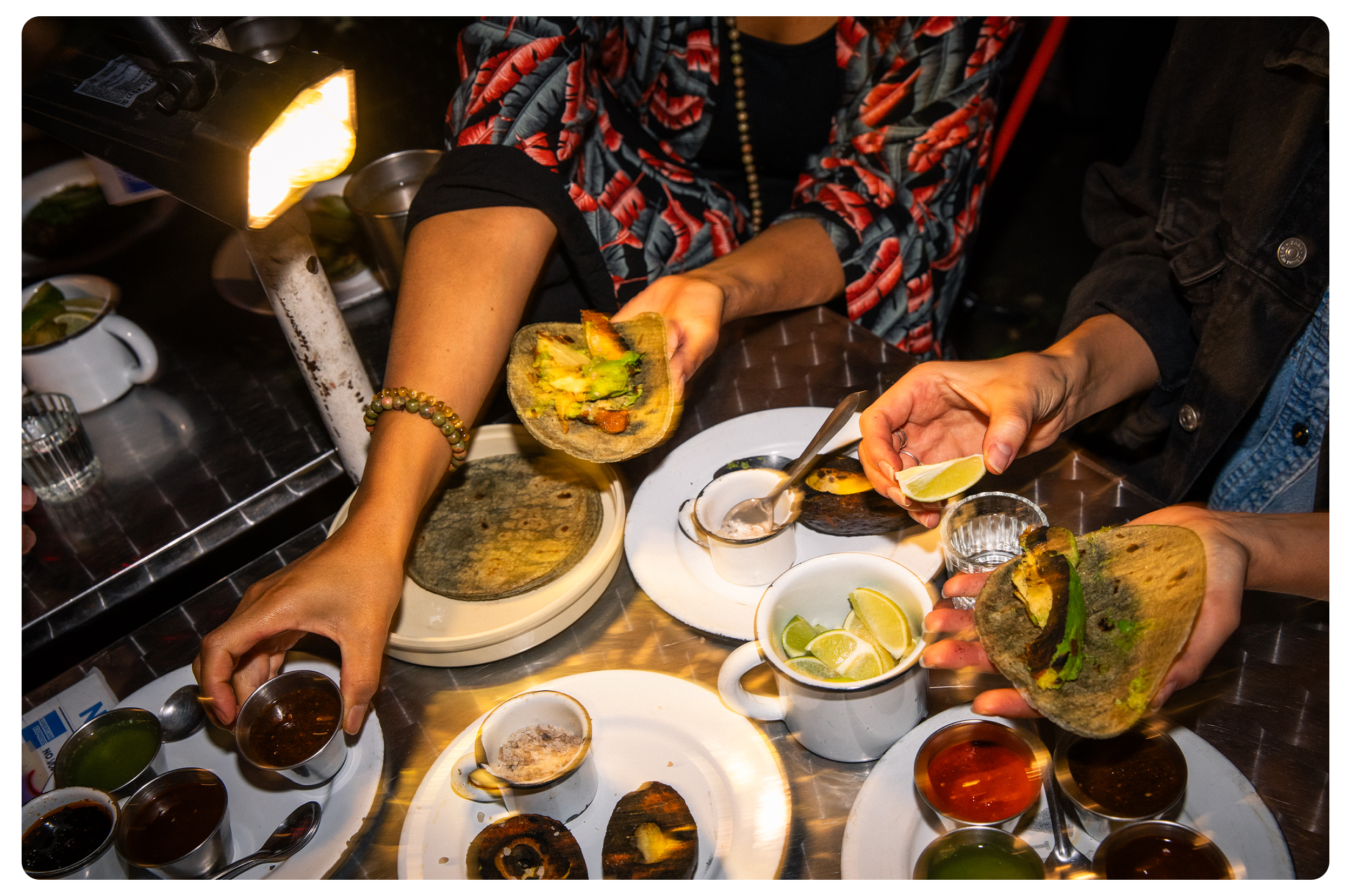Two people dining at a table with Mexican food, including tacos, lime wedges, sauces, and grilled meats, with one person serving tacos and lime.