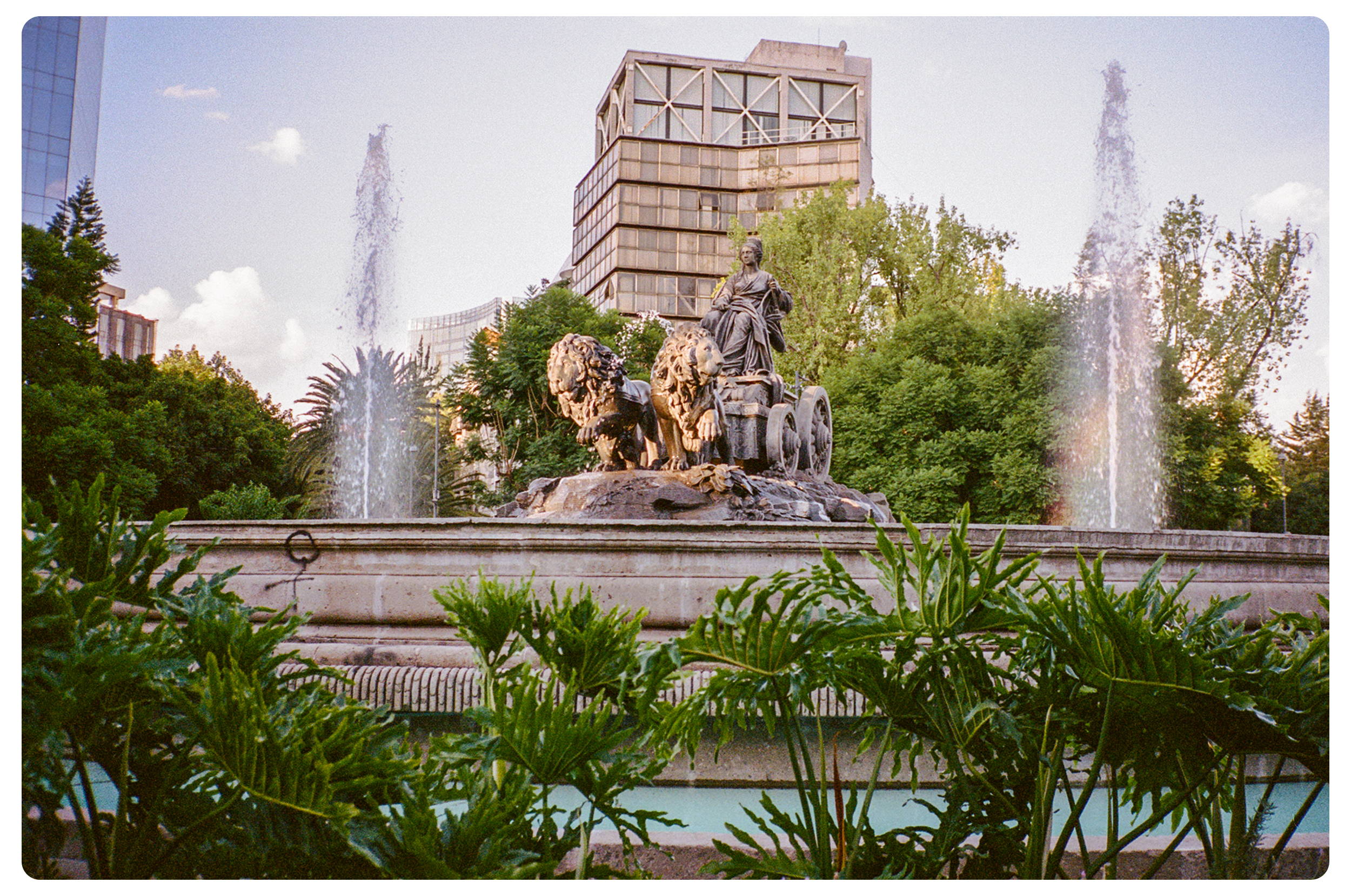 A fountain featuring a statue of a woman riding a chariot pulled by two lions, surrounded by trees and water jets, with a modern building in the background.