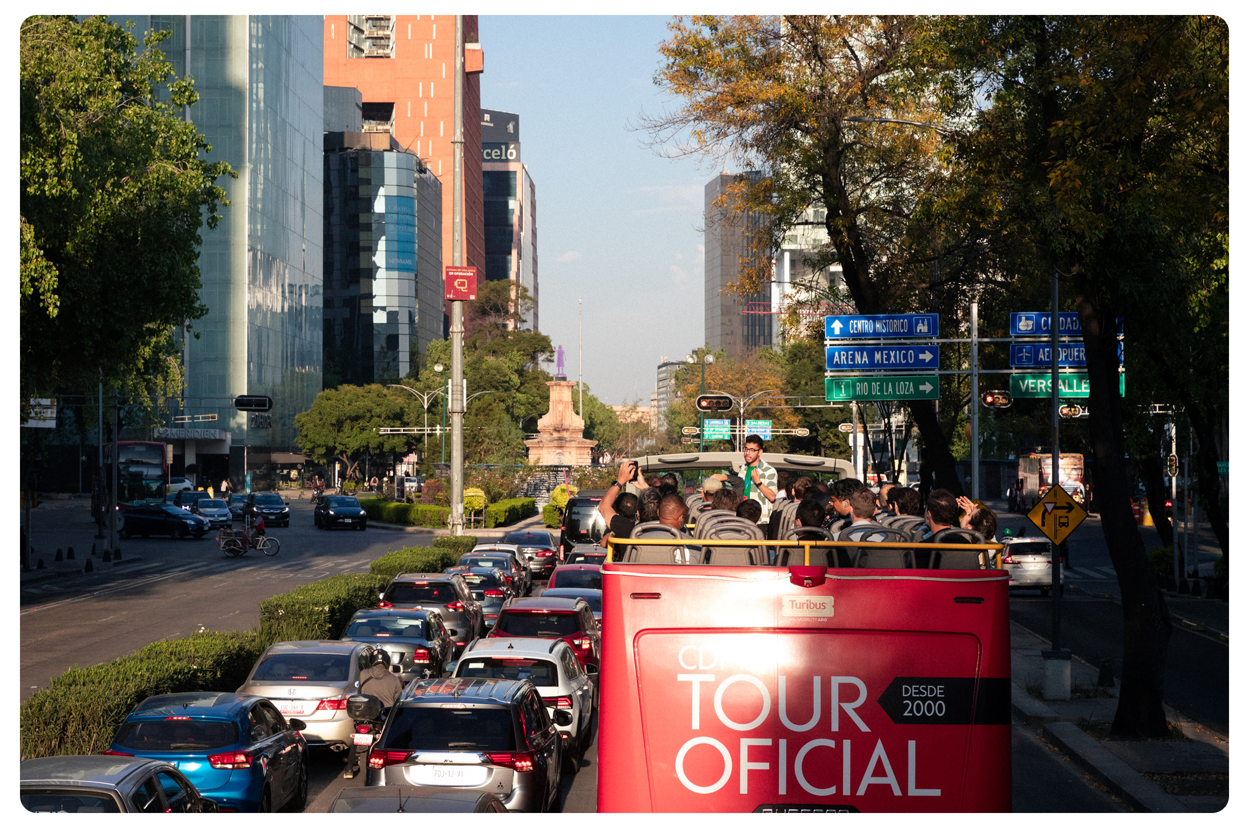 Tourist bus with open roof carrying people in Mexico City, with traffic and high-rise buildings in the background.