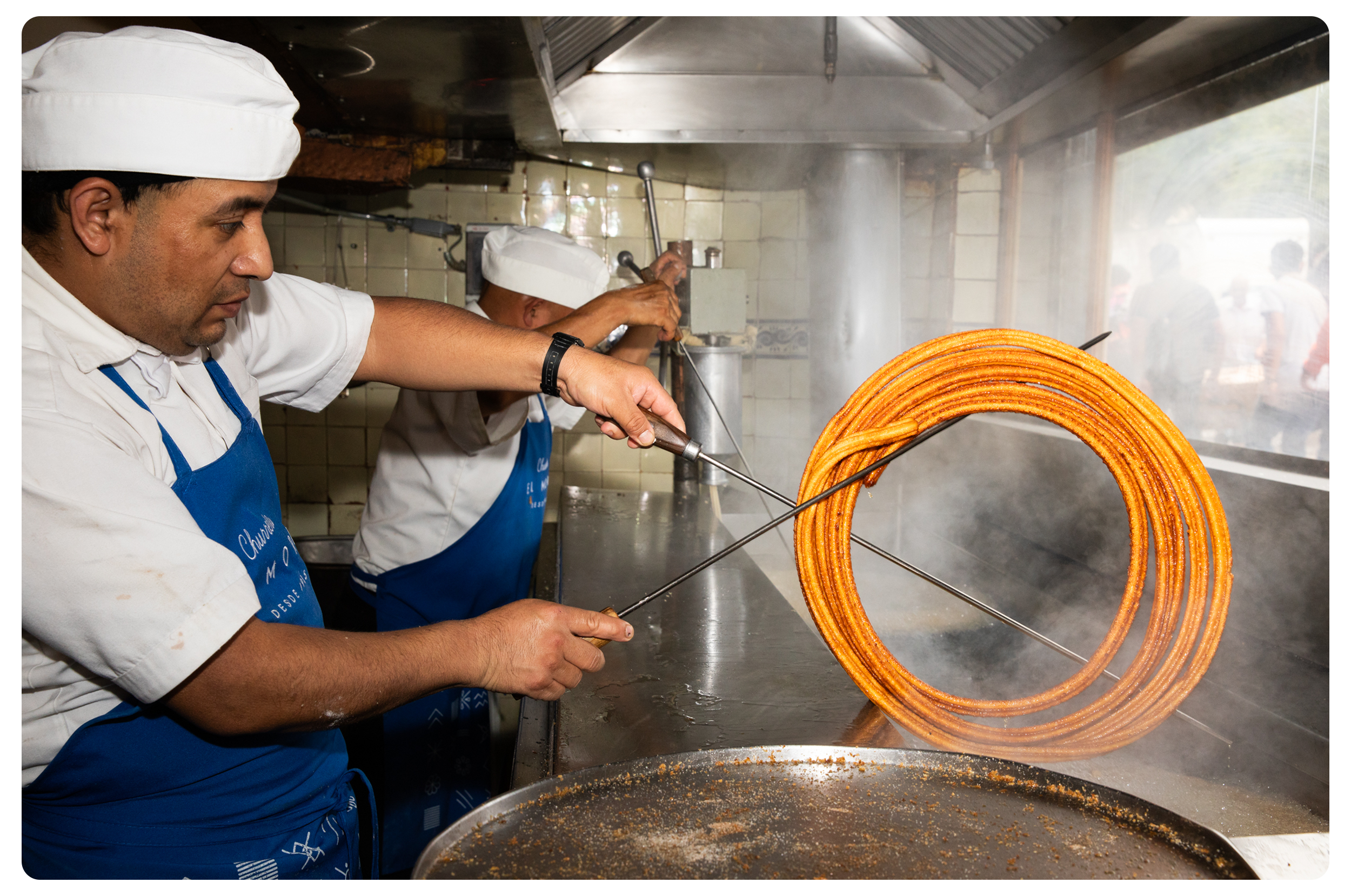 Chef frying churros in large hot oil, using tongs to handle them in a commercial kitchen.