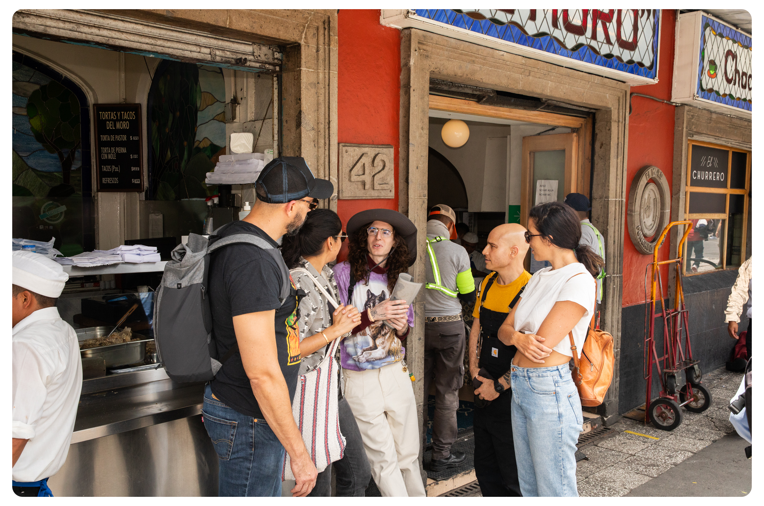 Group of five people standing and talking outside a restaurant entrance on a city sidewalk, with a chef at work inside and a security guard in the background.