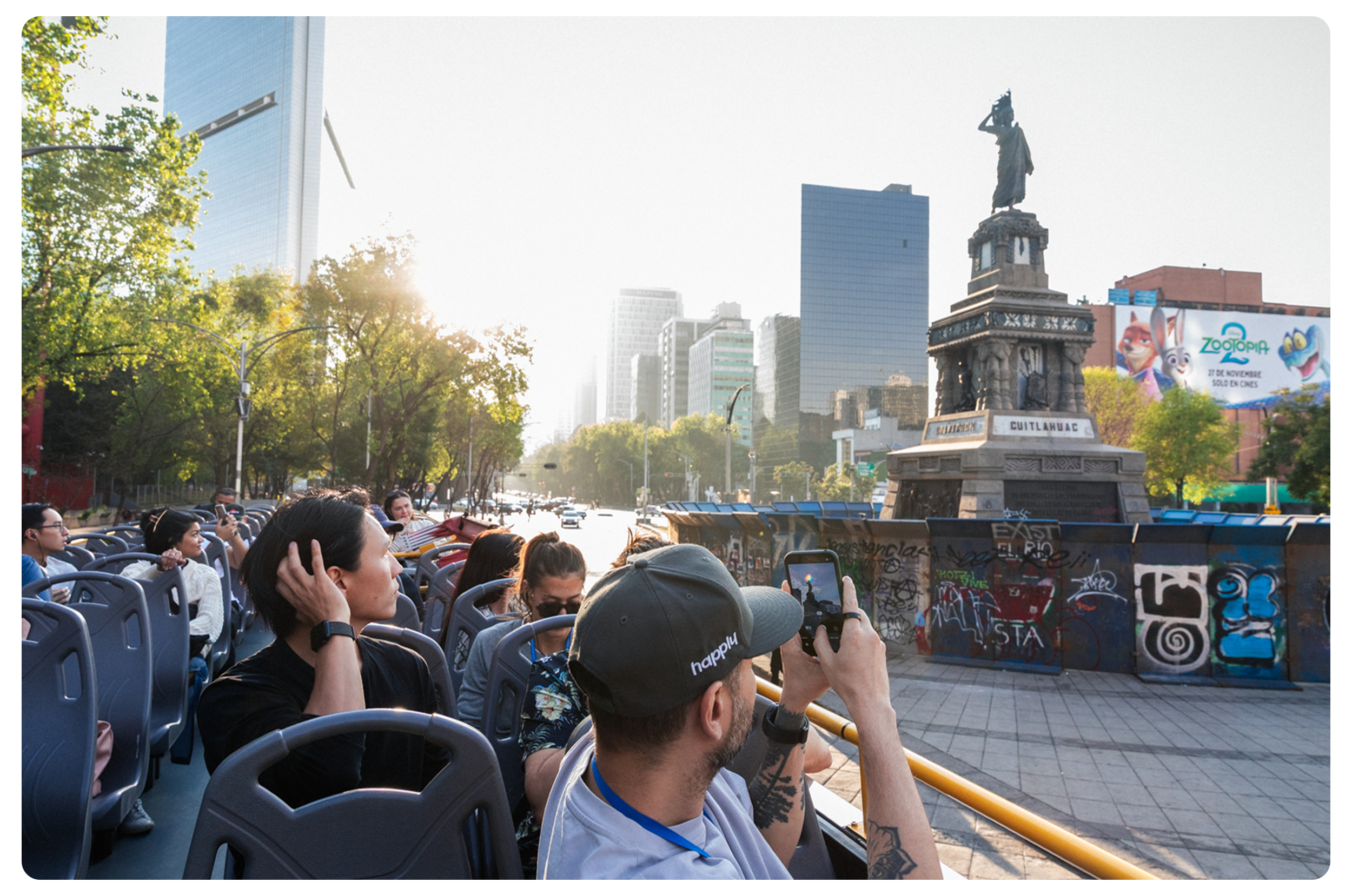 Tourists on a bus sightseeing in Mexico City with the Angel of Independence monument and city skyscrapers in the background.