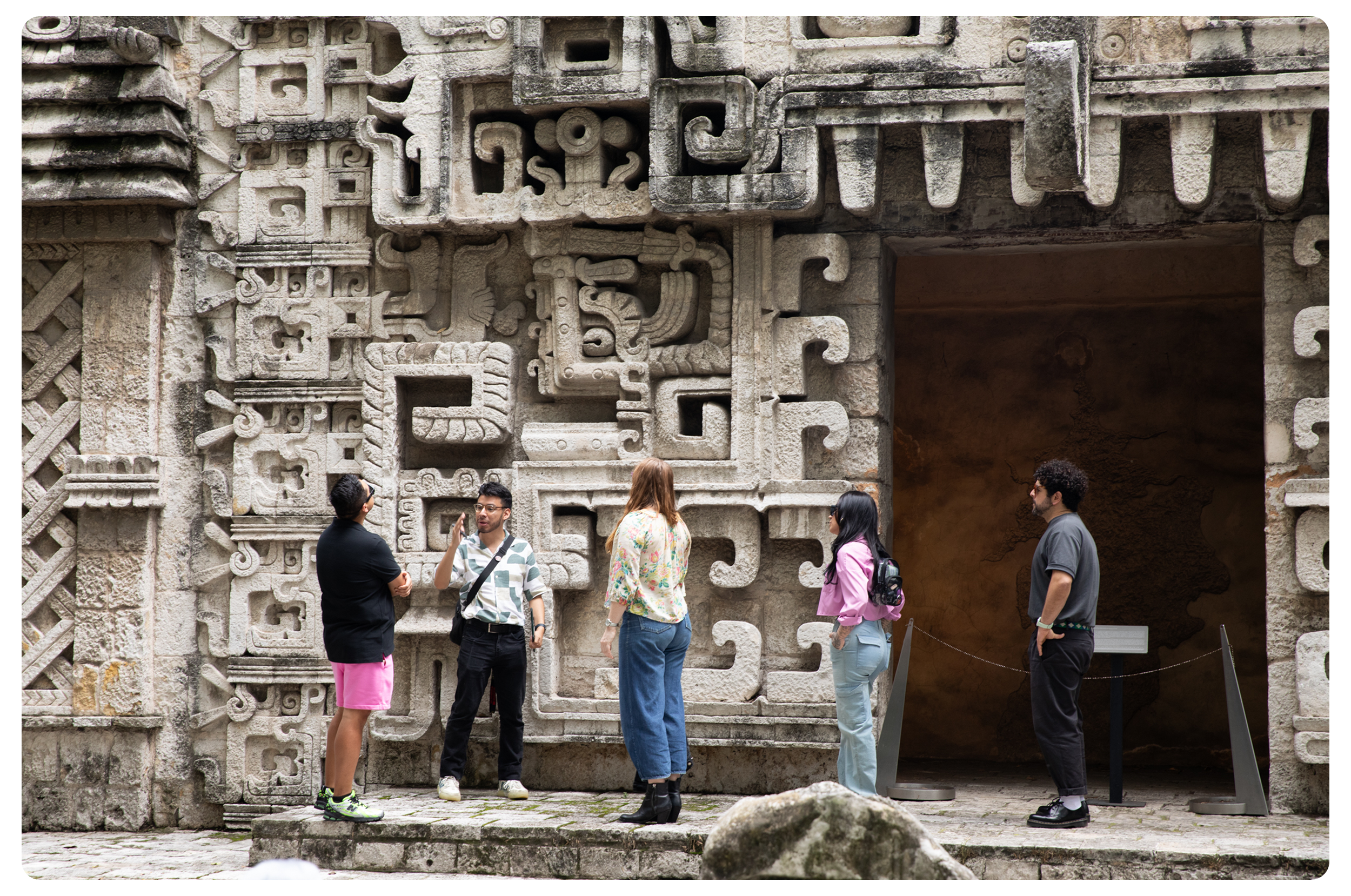 Tourists in front of a carved stone wall at an ancient Mesoamerican archaeological site, engaging in conversation.