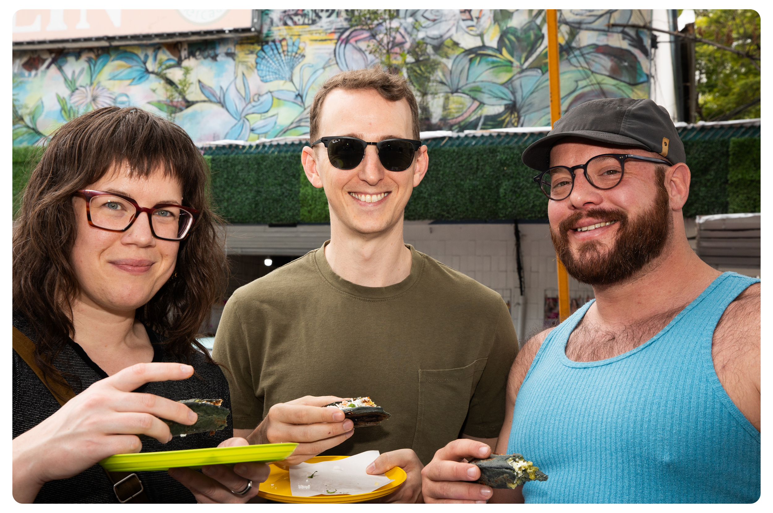 Three people smiling and holding food at an outdoor event, with a mural painted wall behind them.