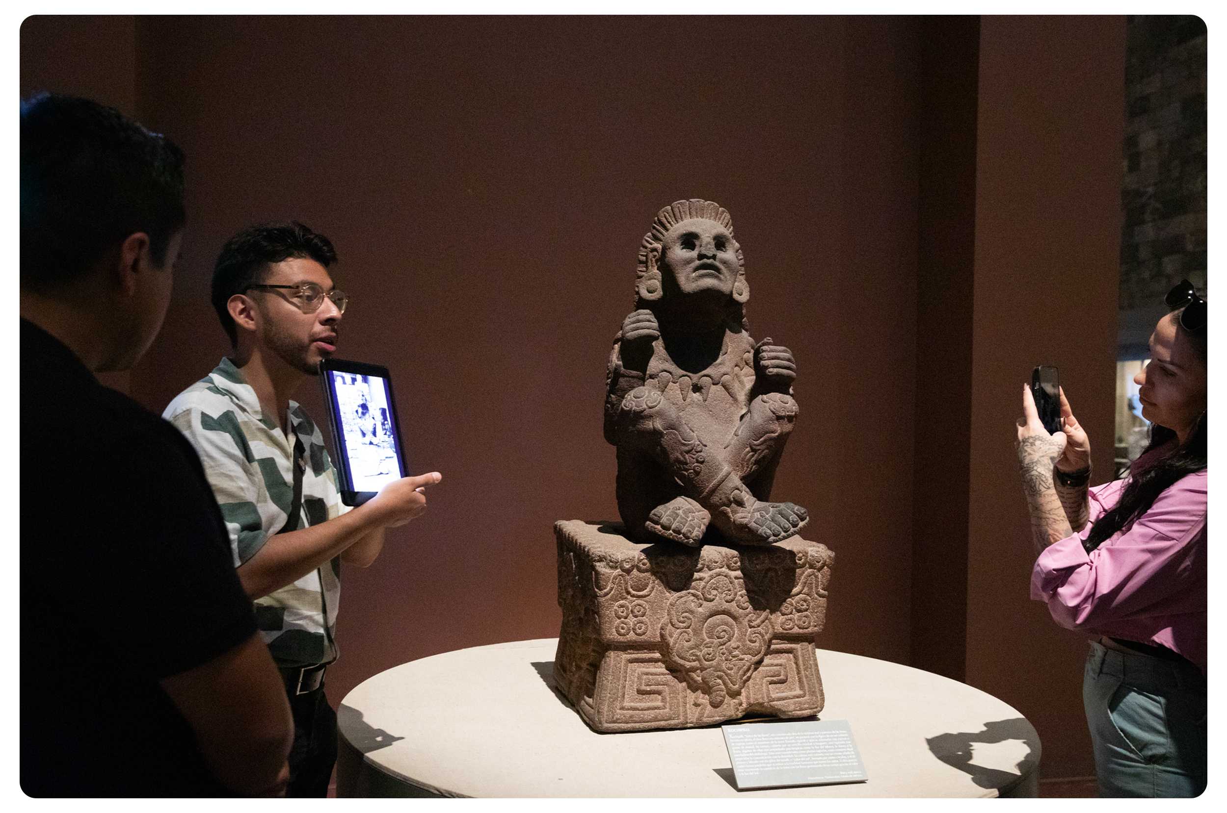 Visitors photographing and viewing an ancient stone sculpture of a seated figure with crossed arms, sitting on a carved pedestal, in a museum setting.
