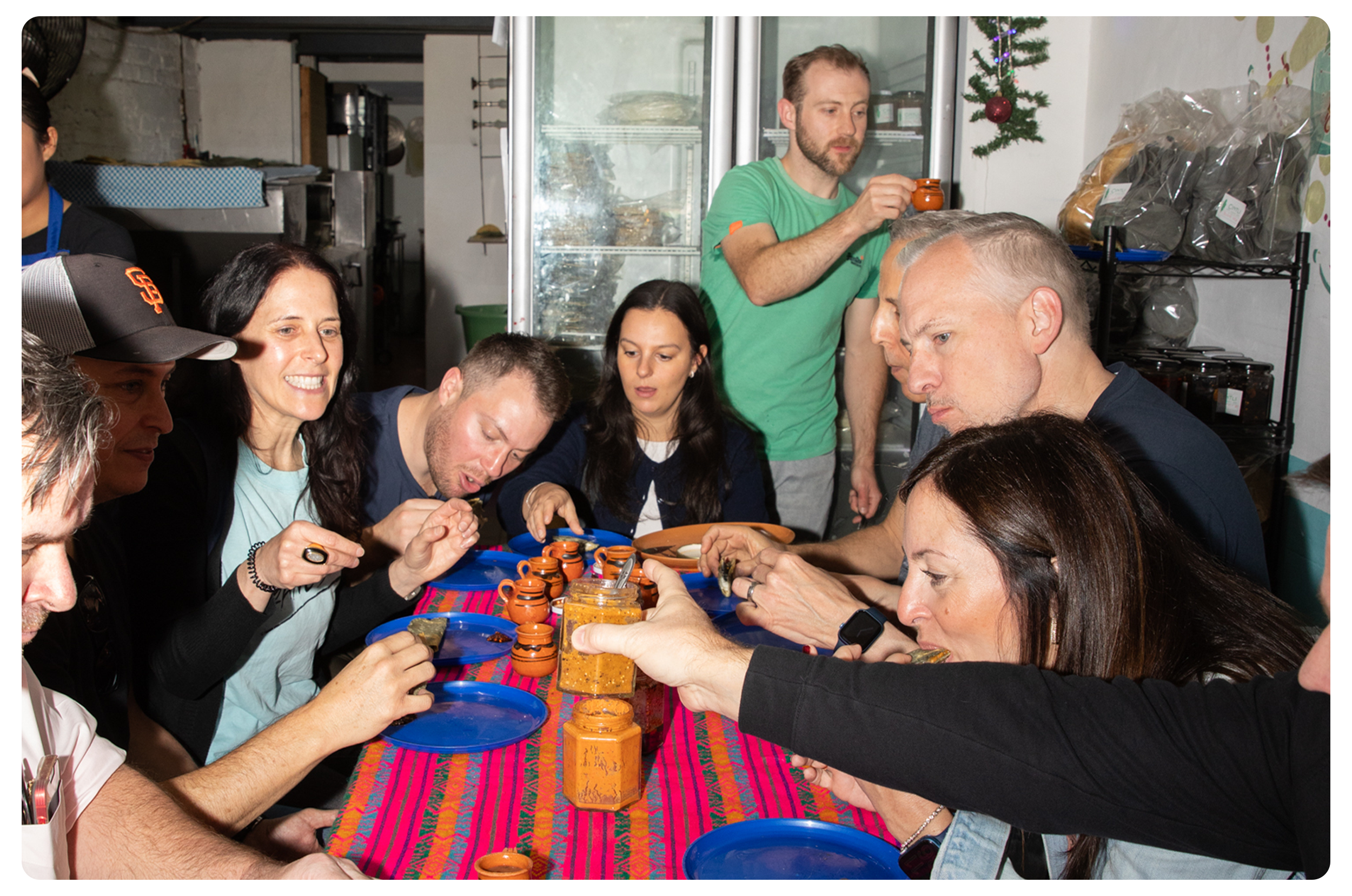 People enjoying Mexican food at a long picnic table in a casual eatery. There are small terracotta sauce containers, jars of salsa, and blue plastic plates on a colorful tablecloth.