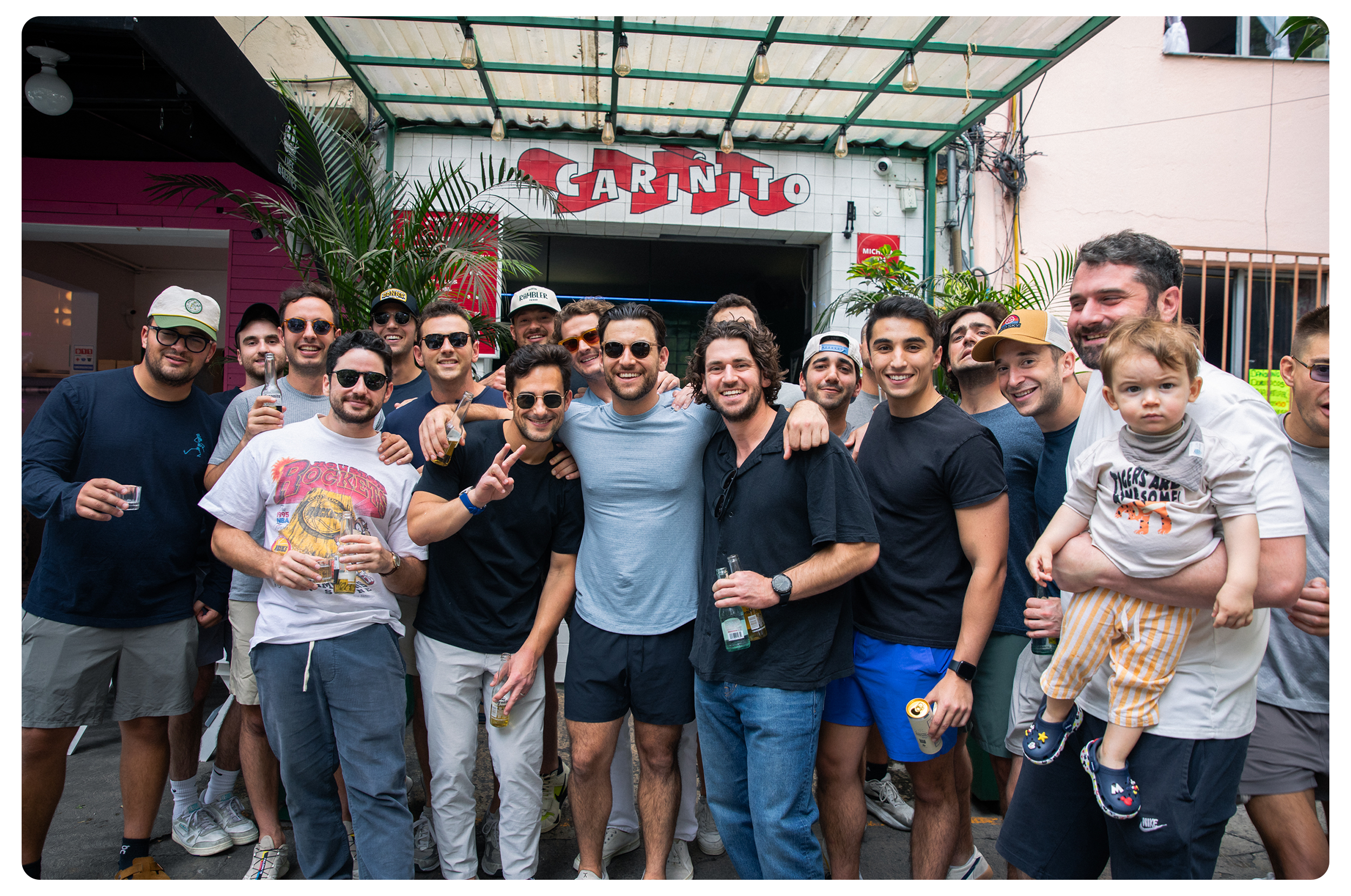Group of friends smiling and posing together outside a bar or restaurant with a sign reading 'CARNITO' in the background. Some are holding drinks, and a man on the right is holding a young child.