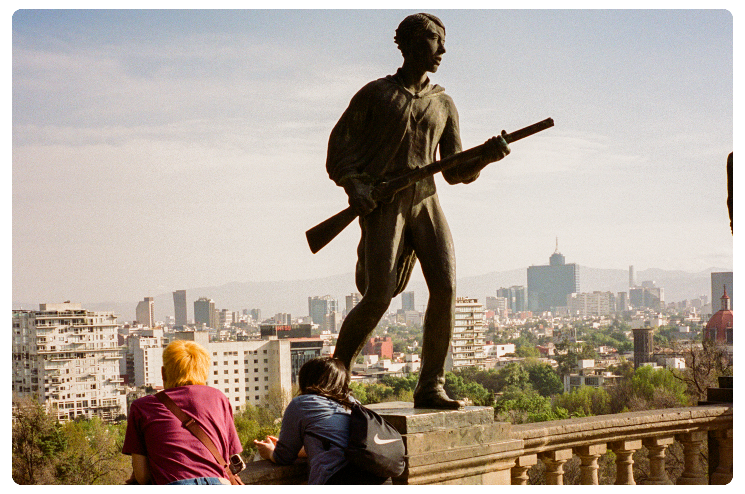 A large bronze statue of a man holding a rifle, located overlooking a city skyline with various buildings, trees, and mountains in the background. Two women are sitting at the base of the statue, enjoying the view.