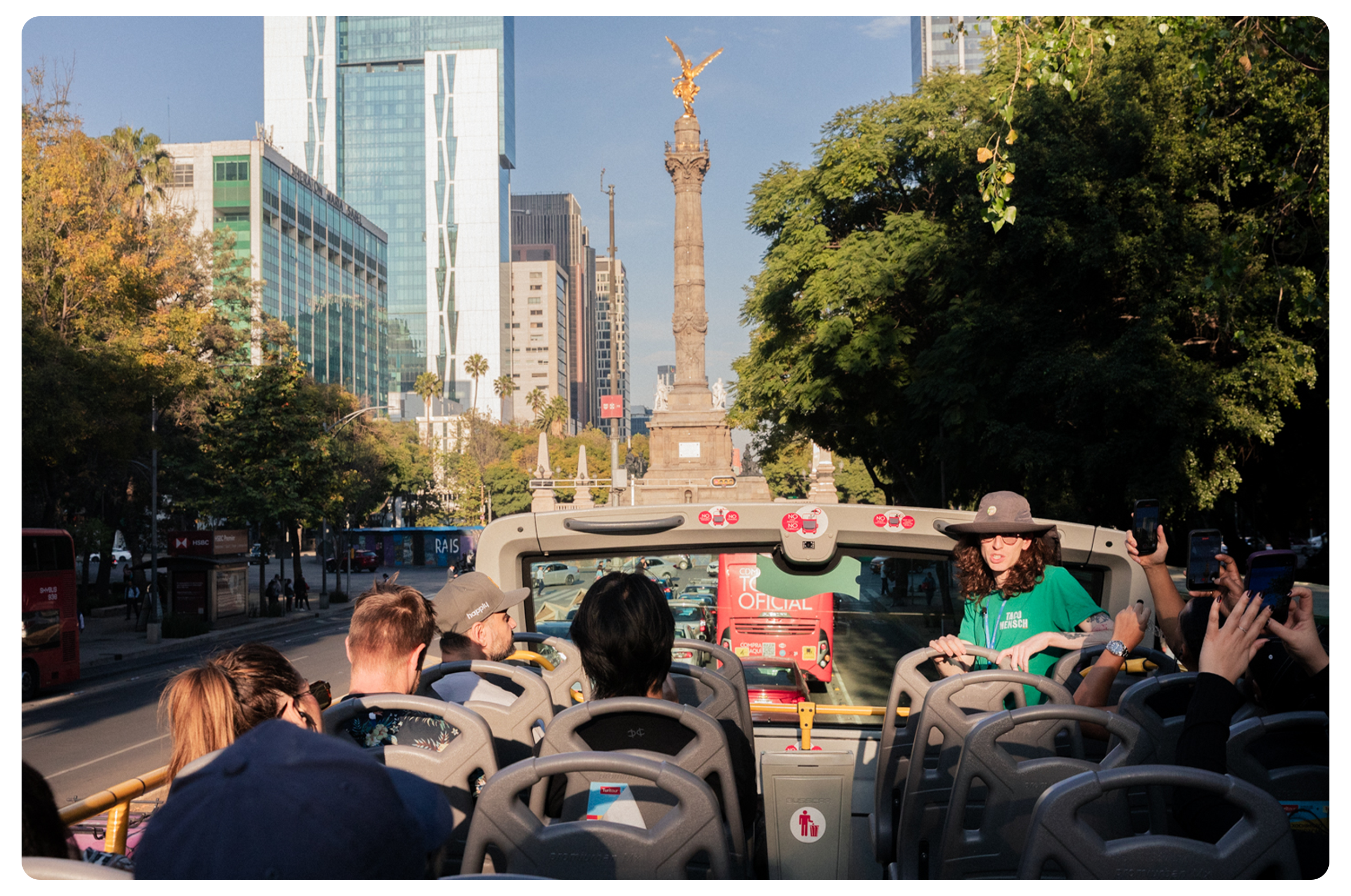 Tourists on open-top bus sightseeing tour in Mexico City with Mexico City National Flag monument in the background, surrounded by tall modern skyscrapers and green trees.