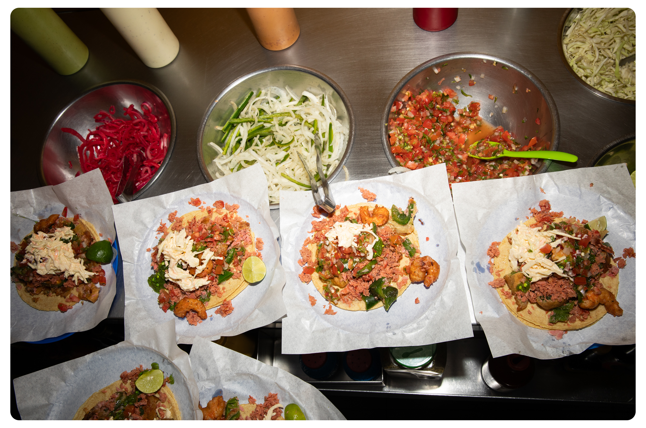 A countertop with four freshly prepared tacos topped with various ingredients, alongside bowls of chopped onions, shredded cabbage, pico de gallo salsa, and red cabbage slaw.