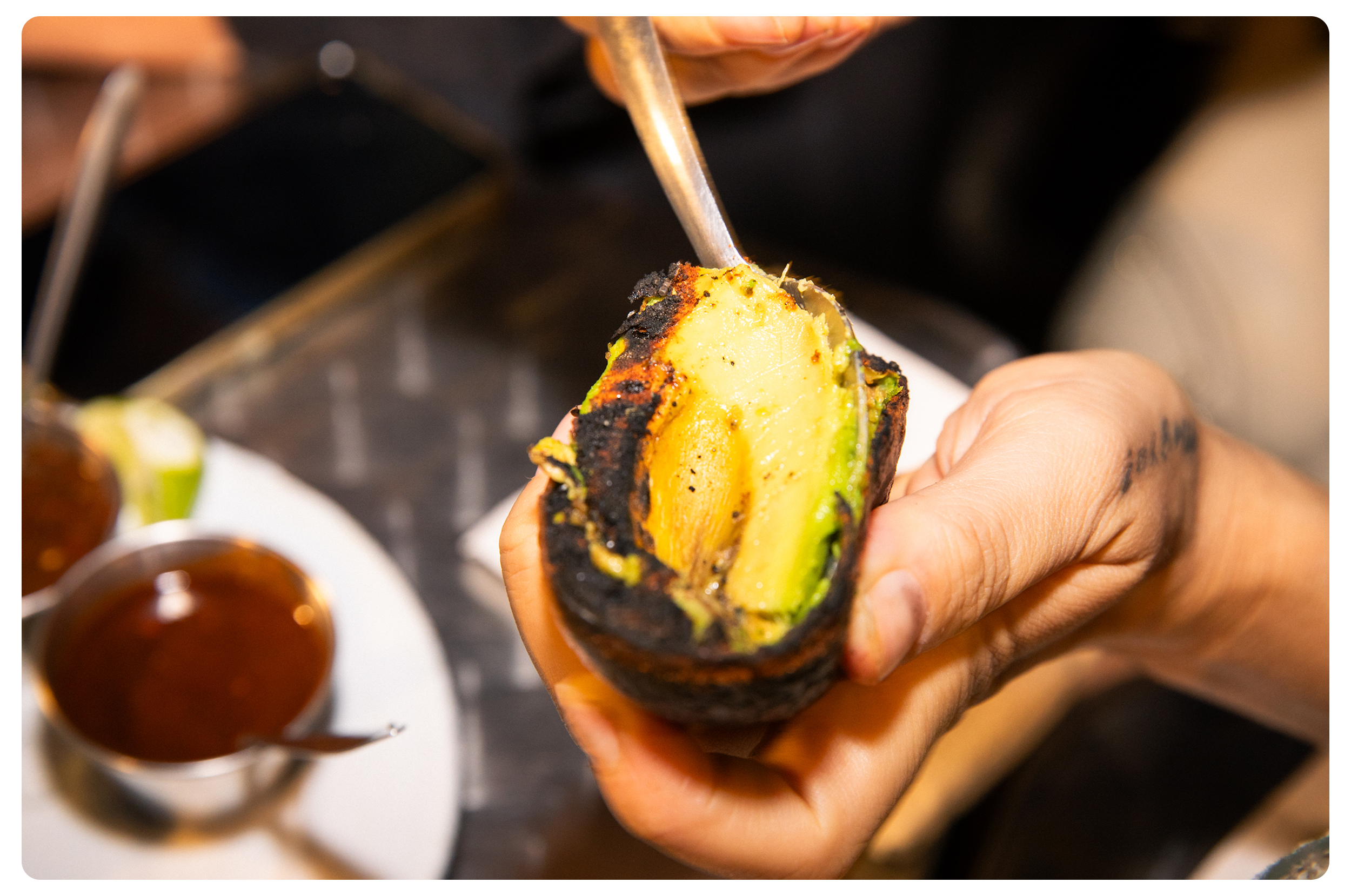 Person holding a grilled avocado with a spoon inside, with a small bowl of sauce or salsa in the background.