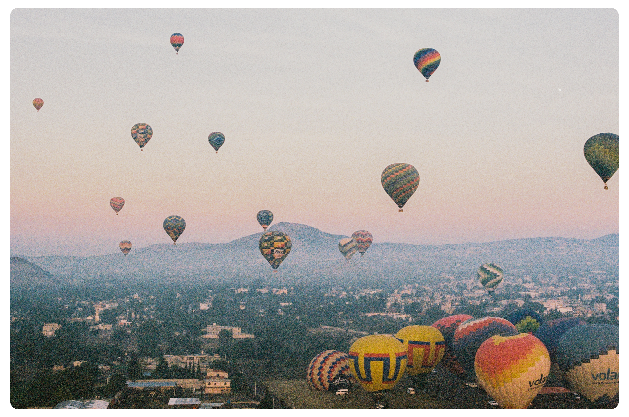 Several colorful hot air balloons floating in the sky above a landscape with hills and buildings at dawn or dusk.