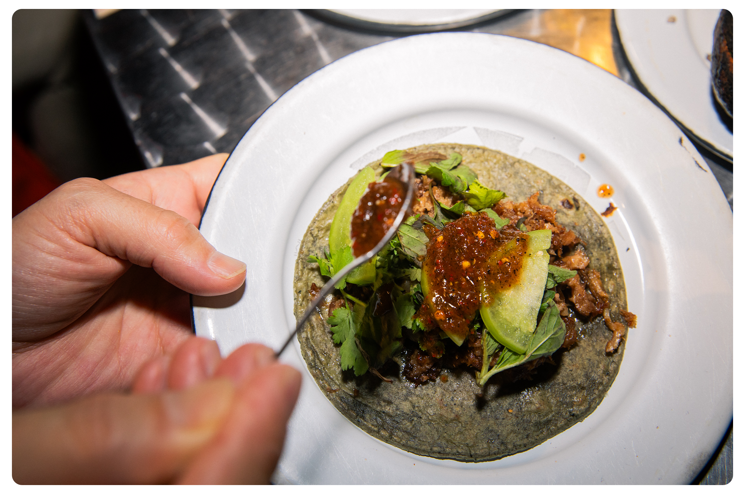 Close-up of a person holding a spoon with red sauce, about to add it to a taco filled with meat, fresh herbs, and lime on a white plate.