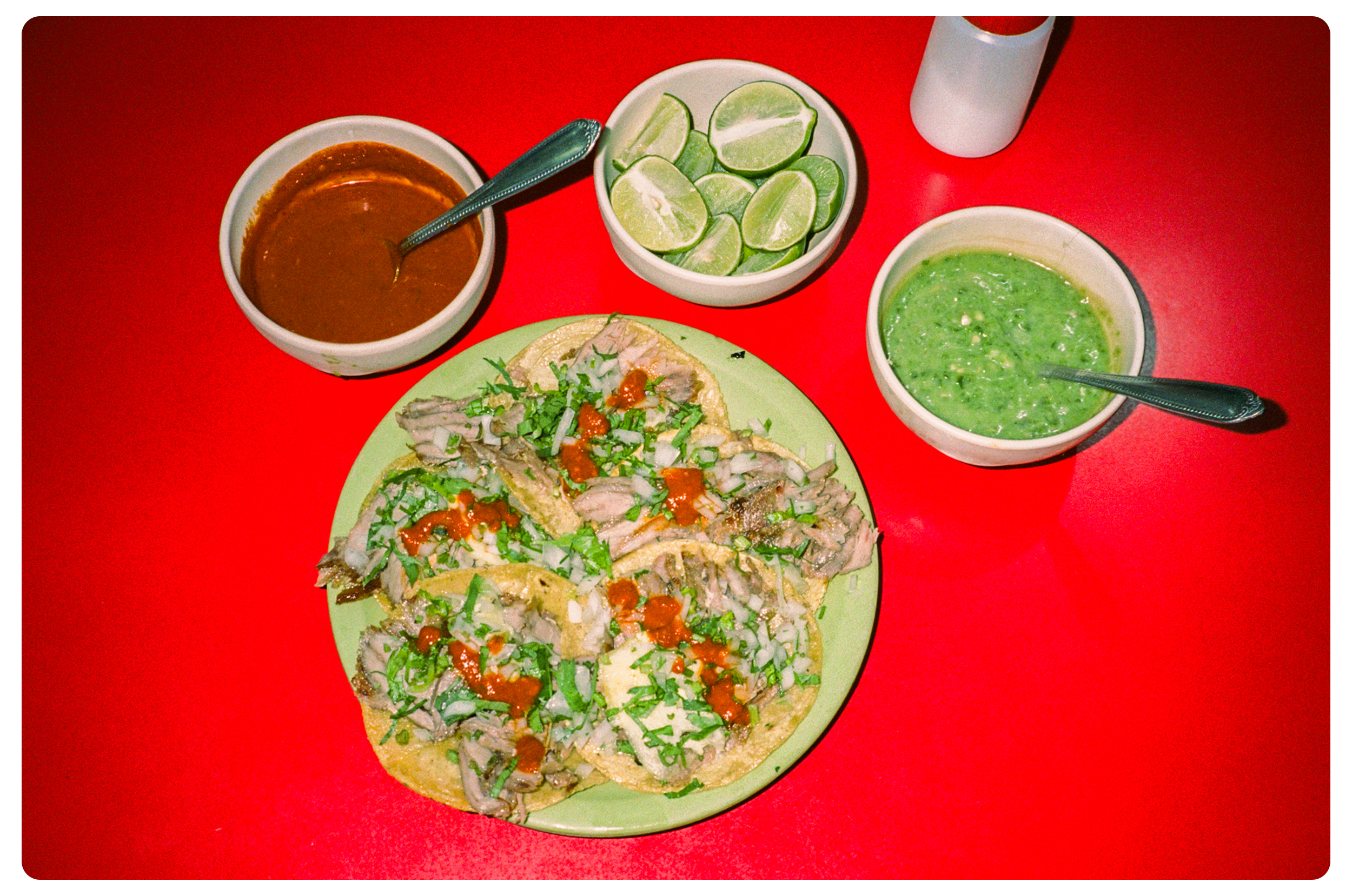 Plate of tacos topped with chopped cilantro, onions, and hot sauce, served with bowls of salsa, guacamole, and lime wedges on a red table.