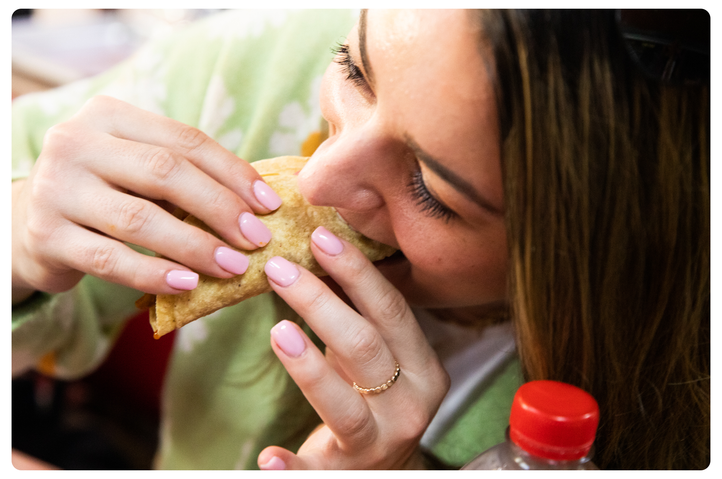 A woman with long brown hair and pink nails is biting into a taco with her eyes closed. There is a water bottle with a red cap near her.