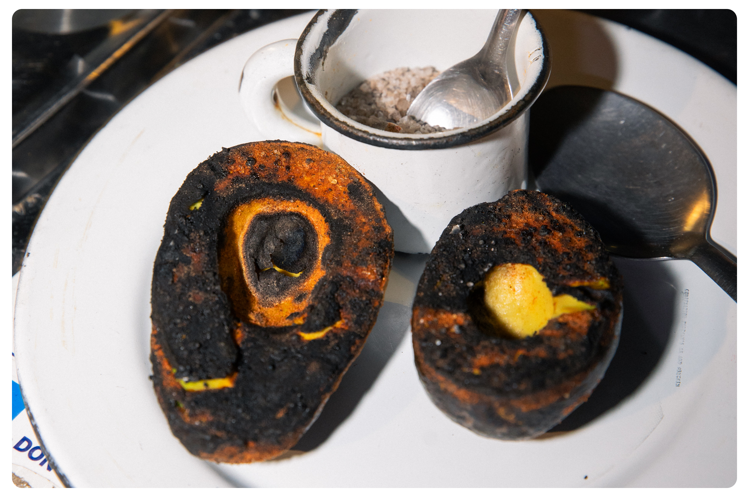 Two roasted plantains with blackened, crispy outer layer on a white plate, with a small container of salt and a spoon nearby.