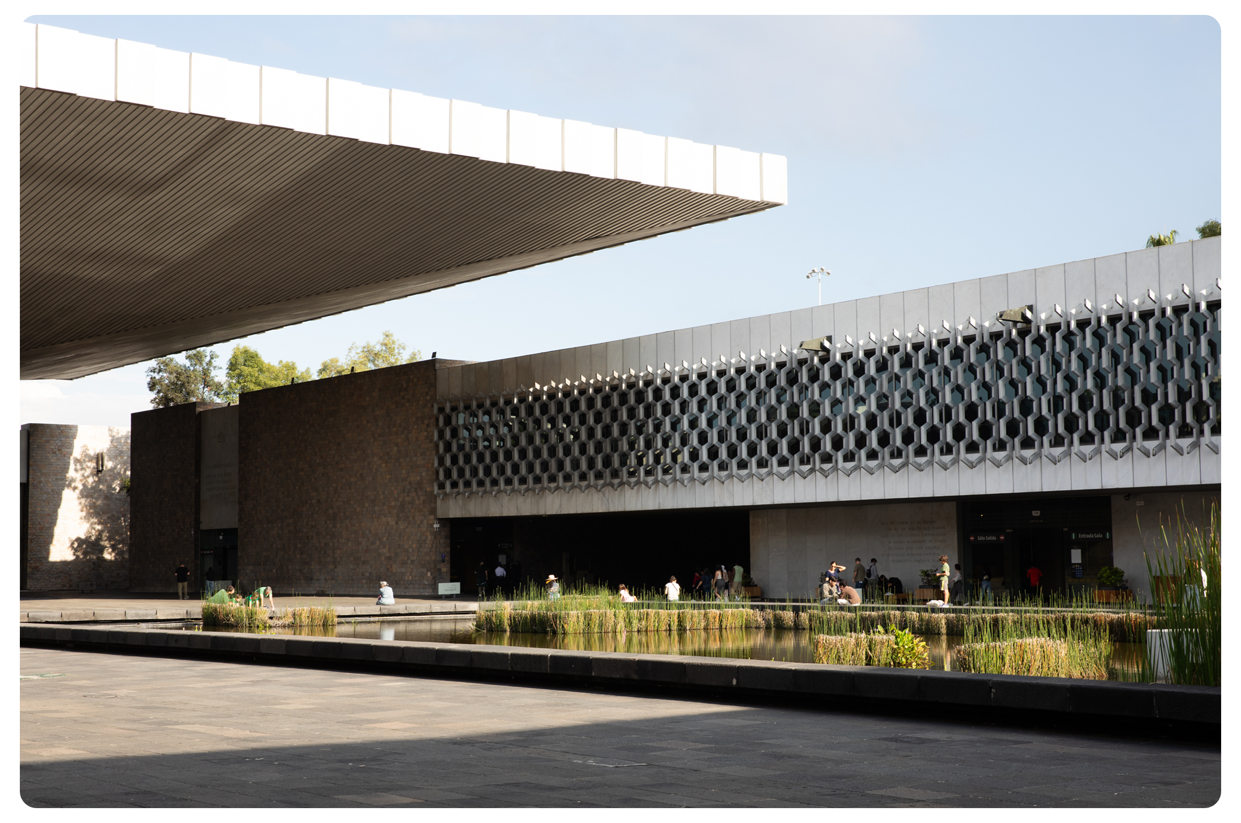 Modern building with geometric patterns, reflecting water and a walkway with people, under a large overhang during daytime.