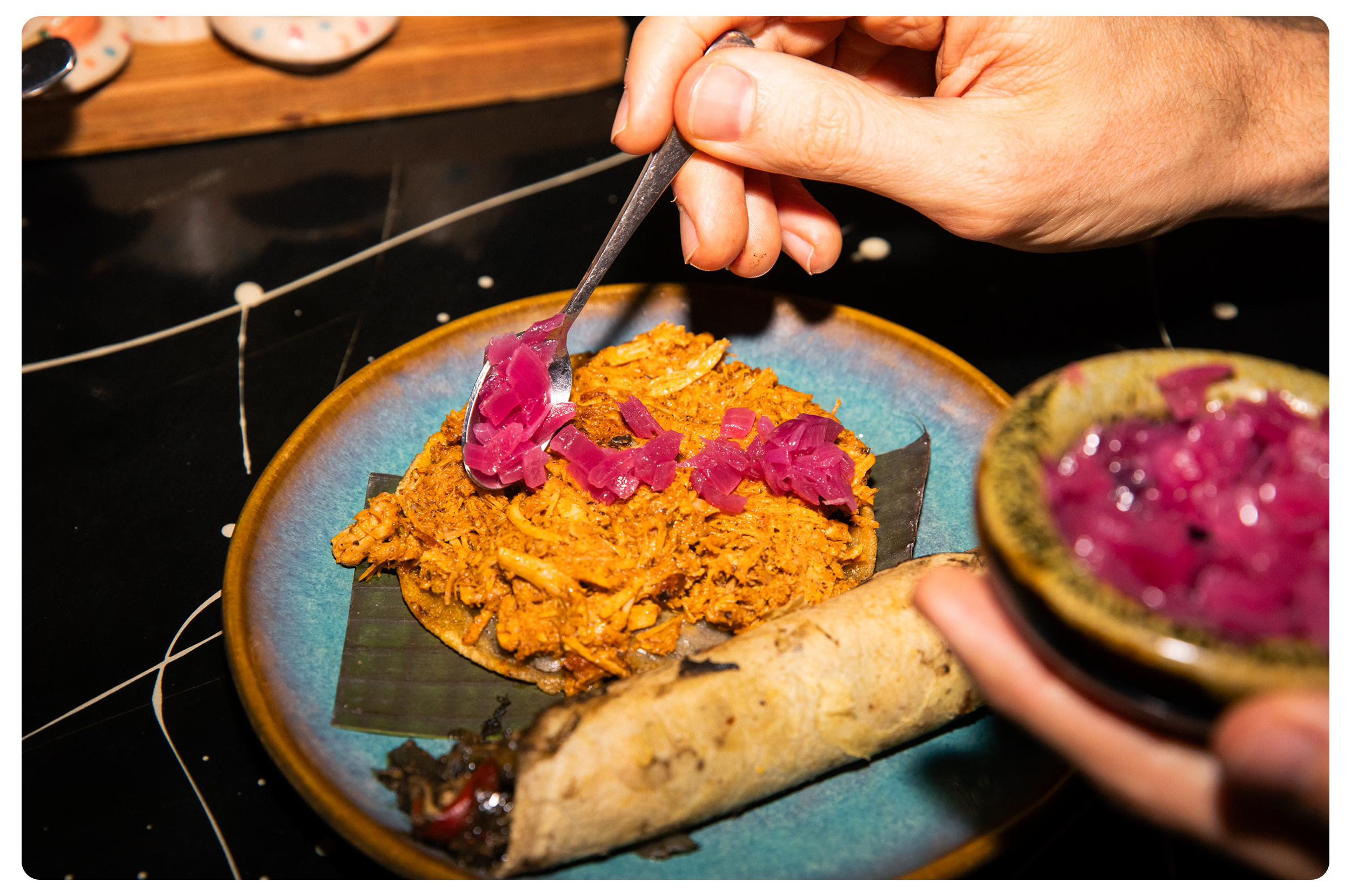 A hand using a spoon to add pickled onions onto a Mexican taco filled with seasoned shredded meat. The taco sits on a blue plate, with a bowl of pickled onions in the foreground.