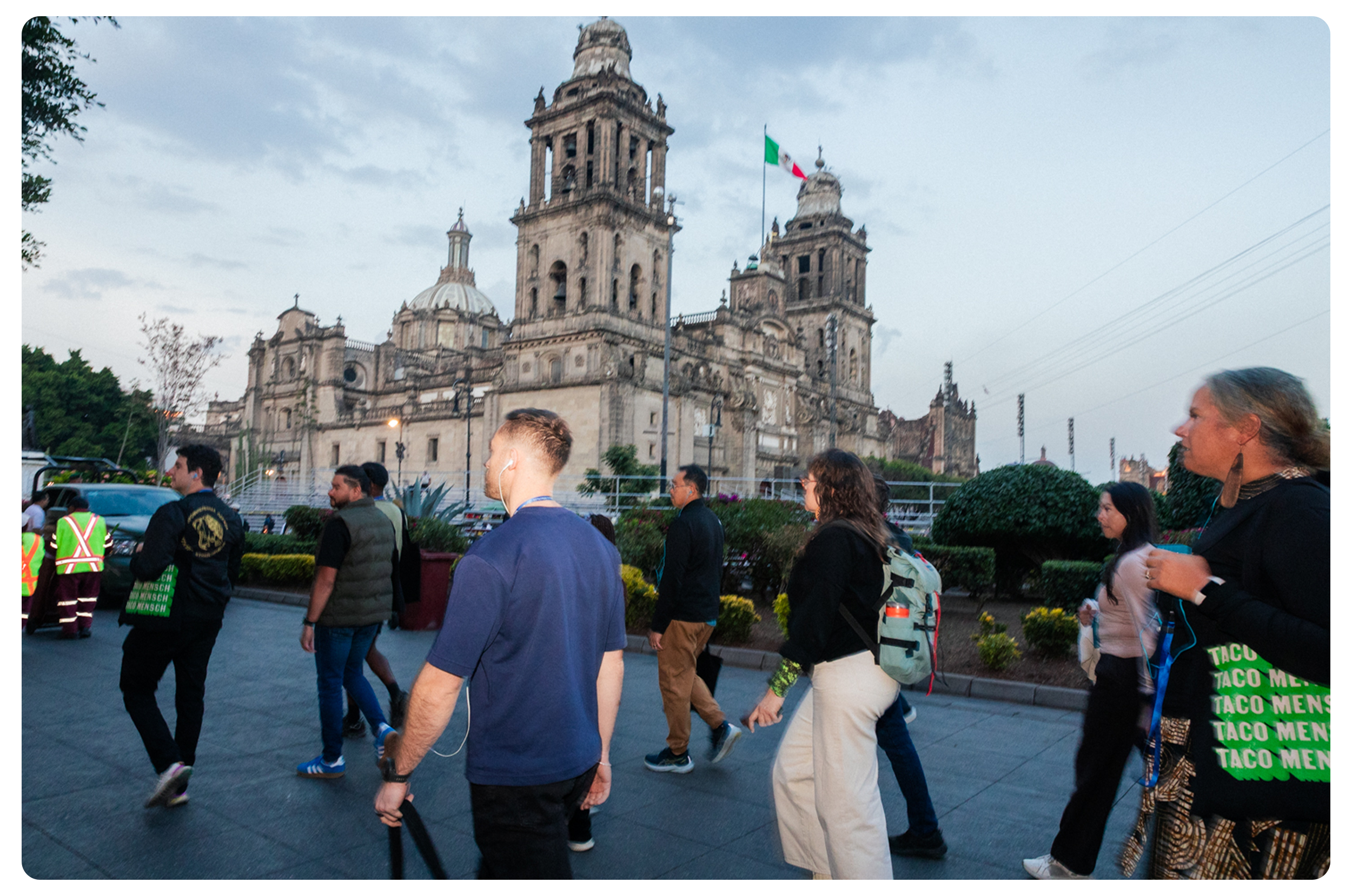 People walking in front of a historic cathedral with two towers, flags, and ornate architecture, late afternoon or early evening.