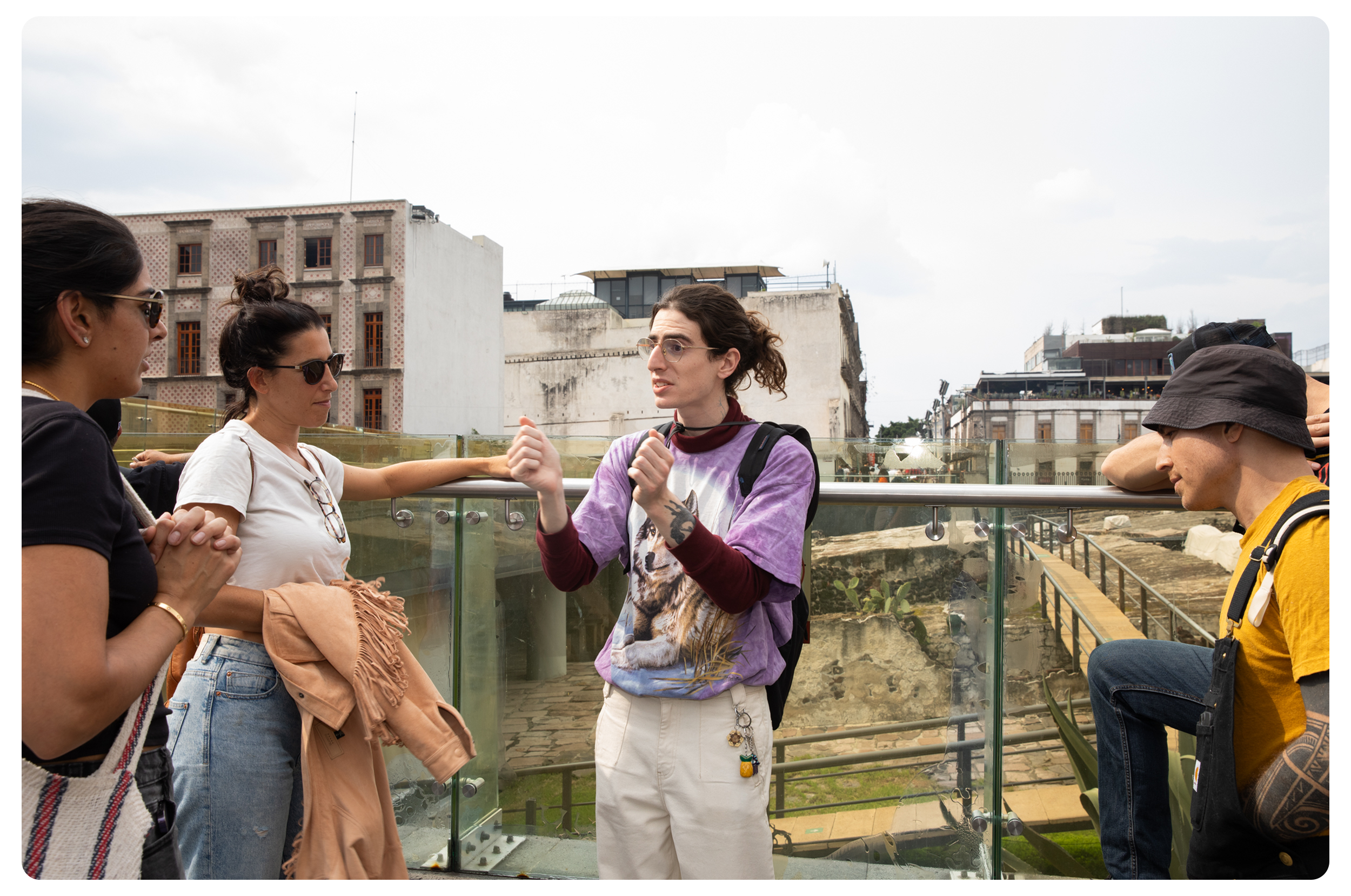A group of people having a discussion on a rooftop with an urban cityscape in the background. One woman is speaking to others, wearing glasses and a colorful graphic t-shirt. Others are listening, wearing casual attire.