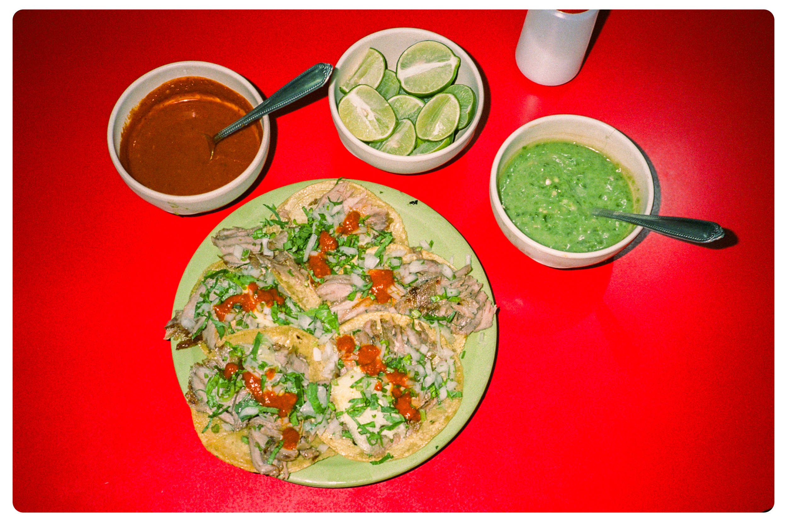 Plate of tacos topped with chopped onions, cilantro, and hot sauce, along with bowls of salsa, guacamole, and lime wedges on a red table.