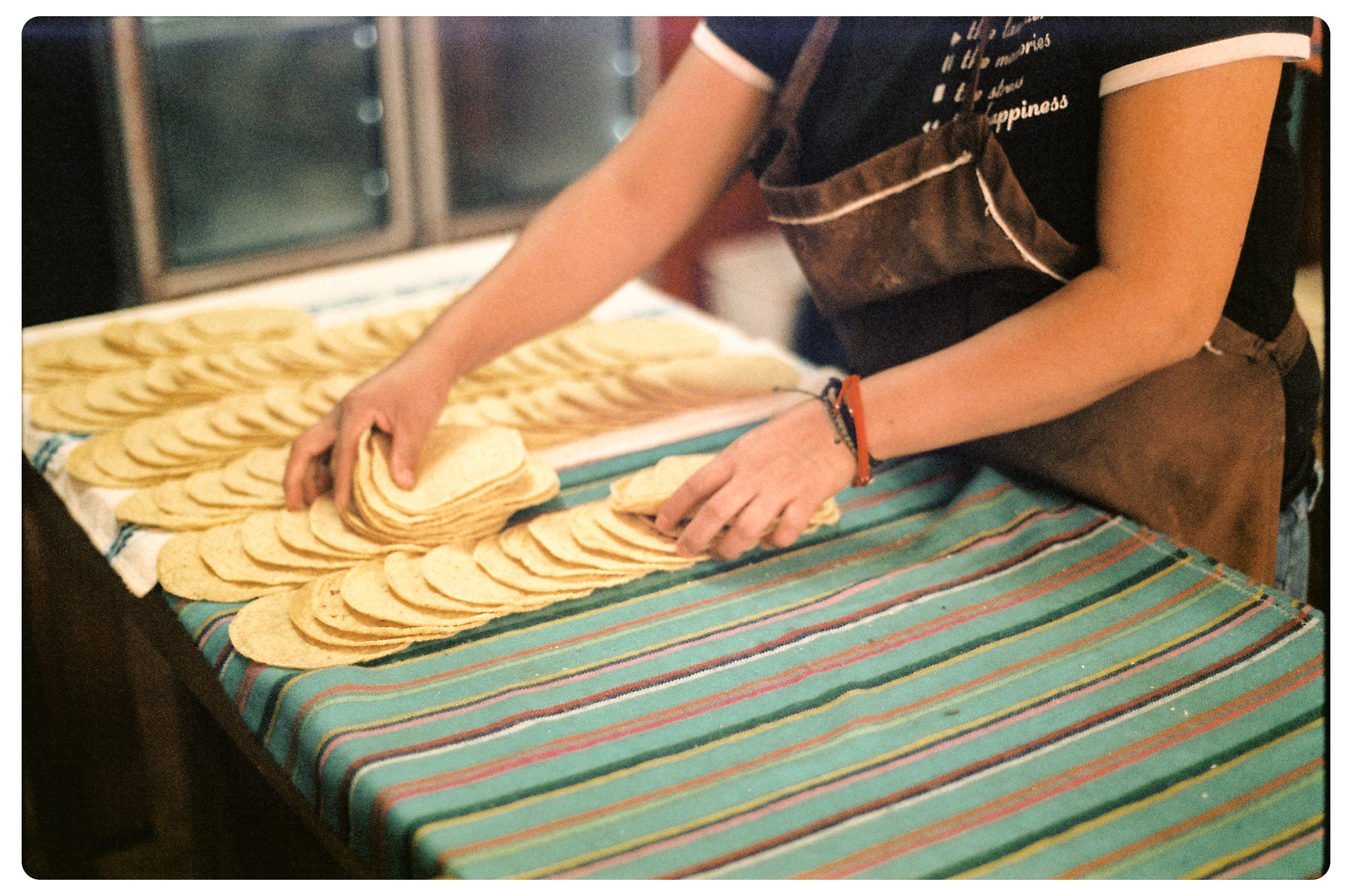 Person wearing a brown apron and black shirt arranging or stacking tortillas on a striped cloth-covered table.