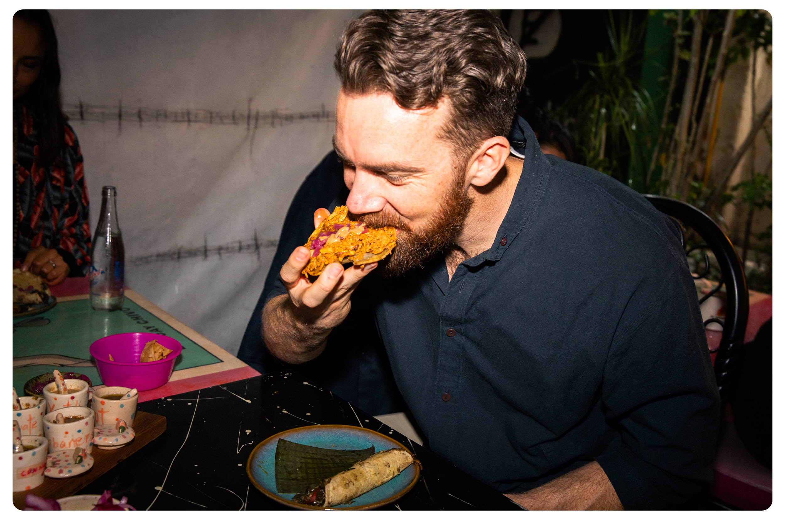 A man with a beard and wearing a dark shirt is taking a bite of a large piece of food, possibly a sandwich or taco, at a table with various dishes and small cups. There are other people and plants visible in the background.