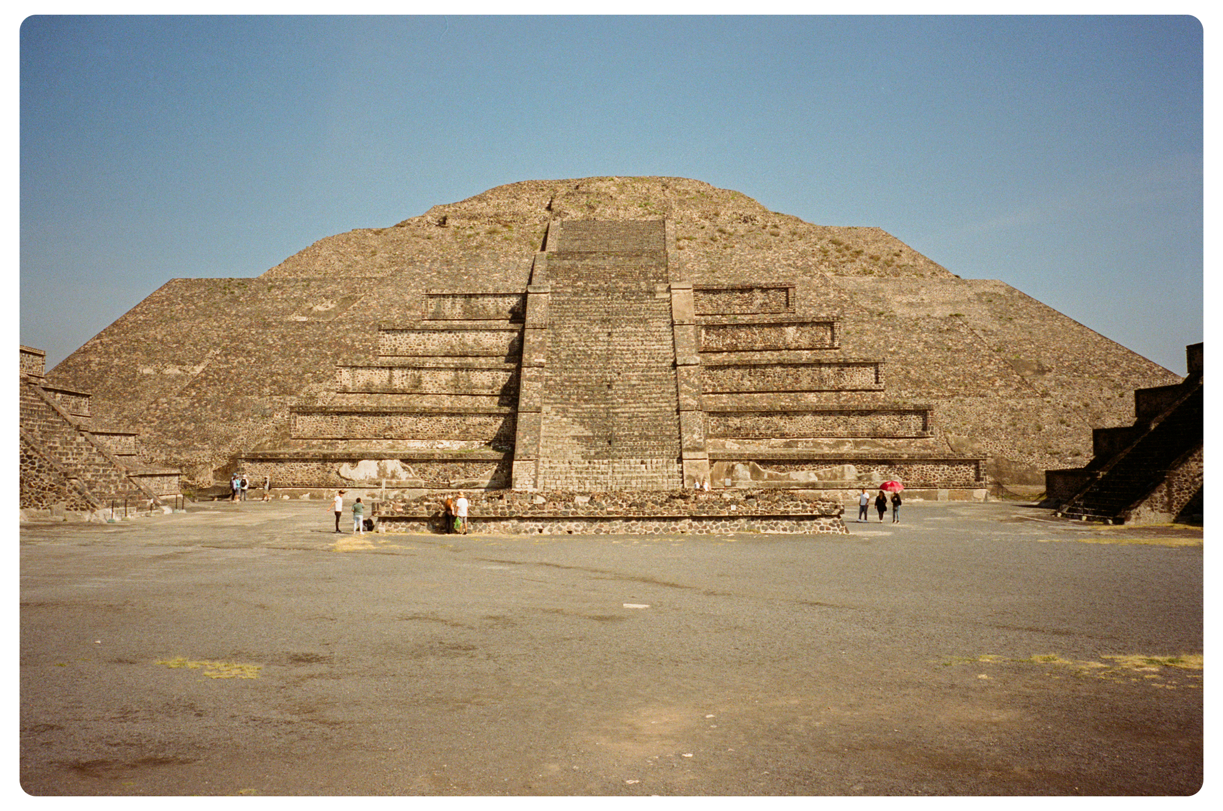 The Pyramid of the Magician at Uxmal in Mexico with a large, stepped pyramid structure, and a flat open plaza in front with a few tourists walking around.