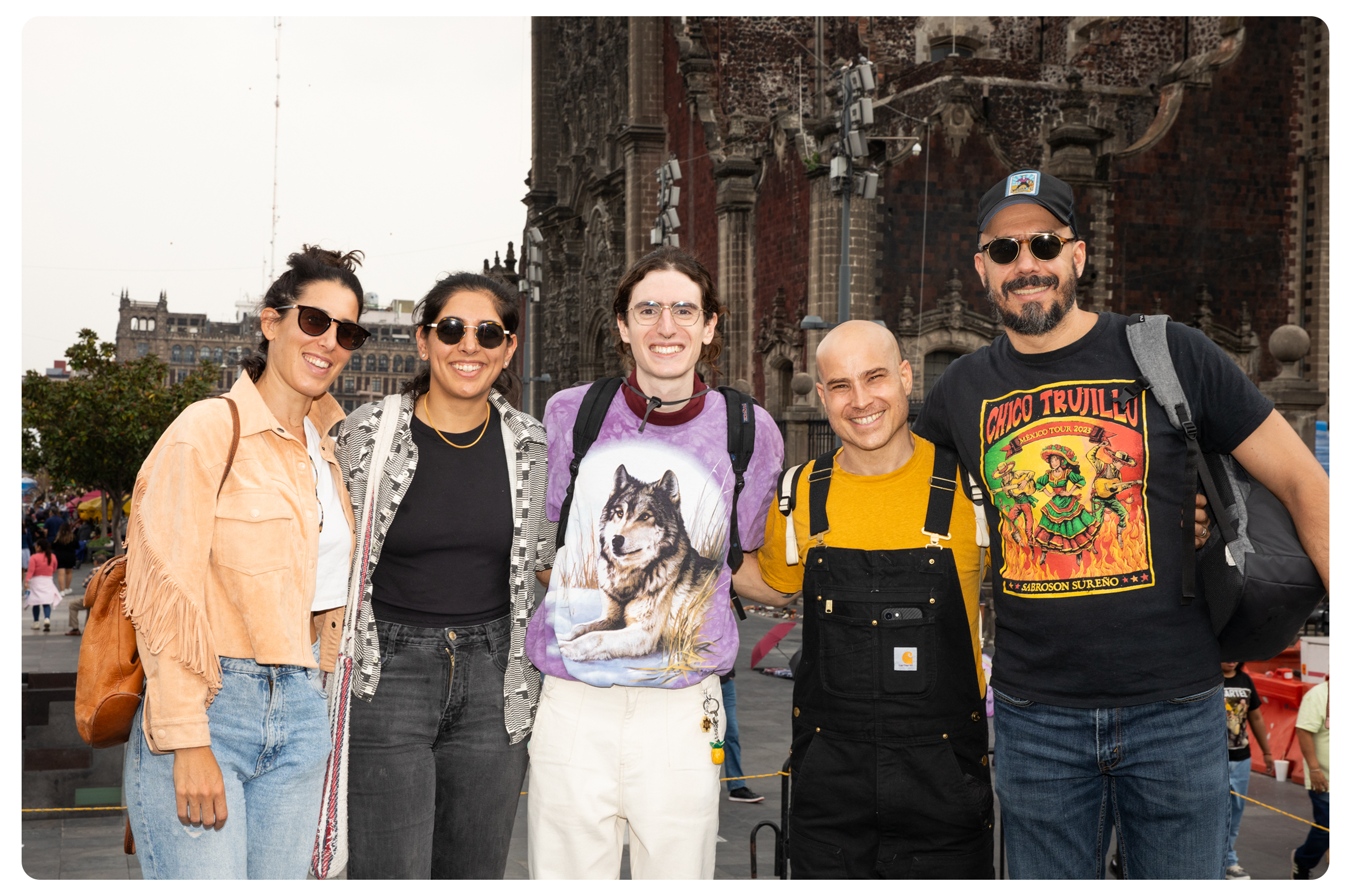 Group of six diverse young adults smiling and posing for a photo outdoors in front of historic architecture, with some wearing sunglasses and casual clothing.