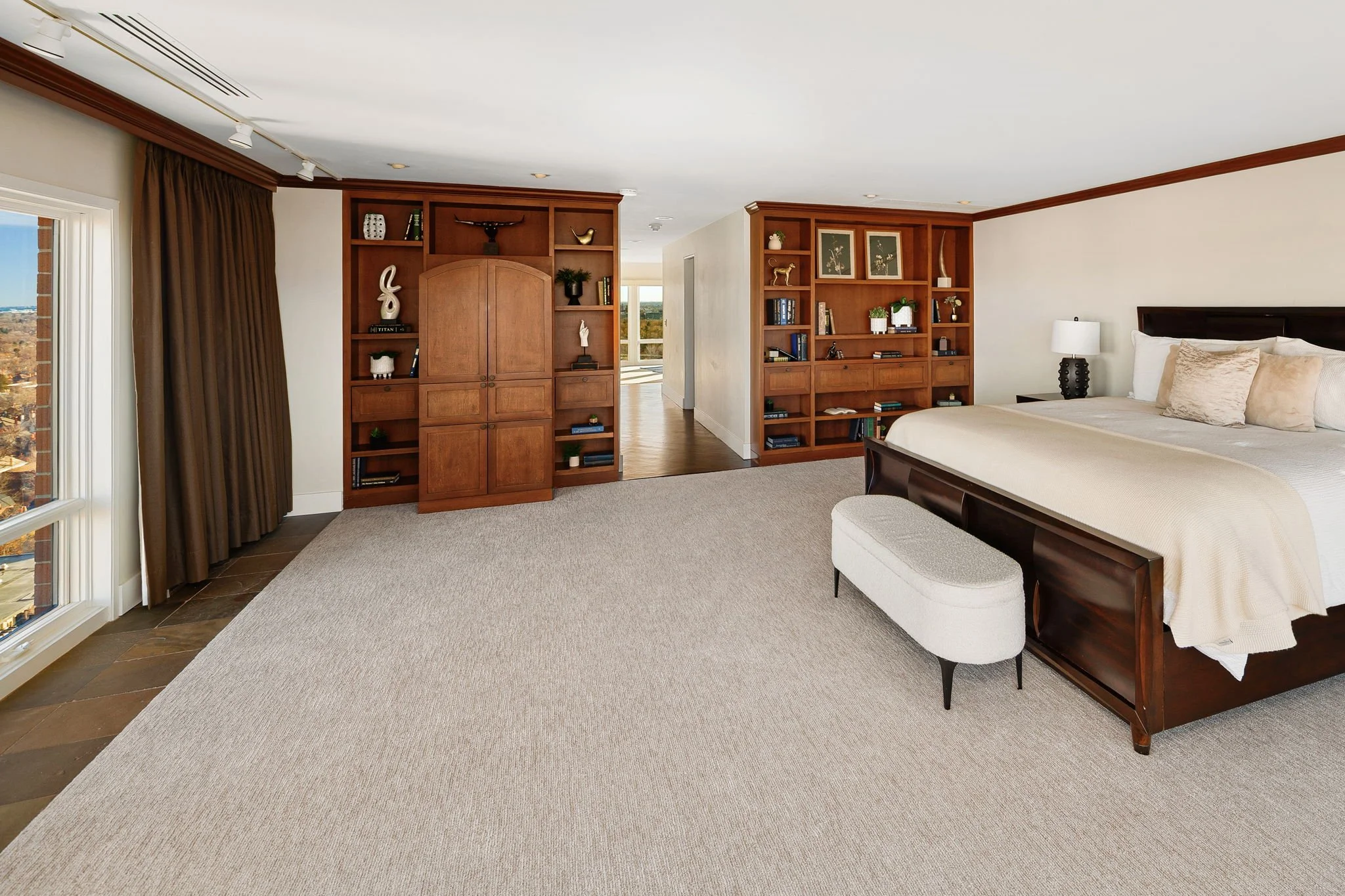 A bedroom with a large bed, nightstand, table lamp, bookshelf, and window with curtains.