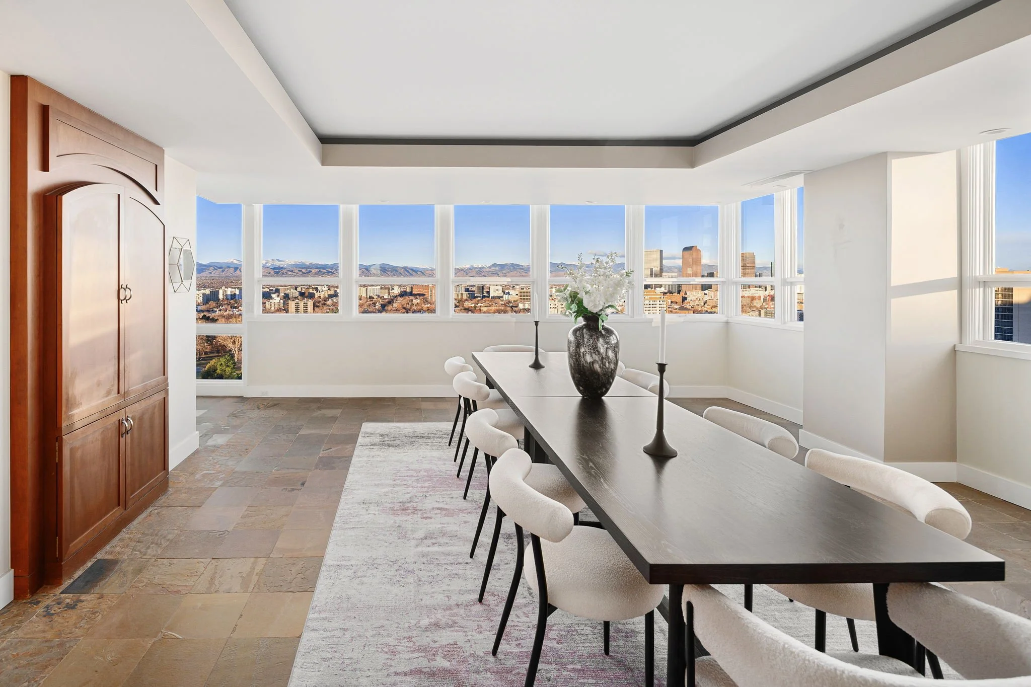 Modern dining room with a long black table, white upholstered chairs, a large vase with white flowers, black candlesticks, and panoramic city and mountain views through large windows.