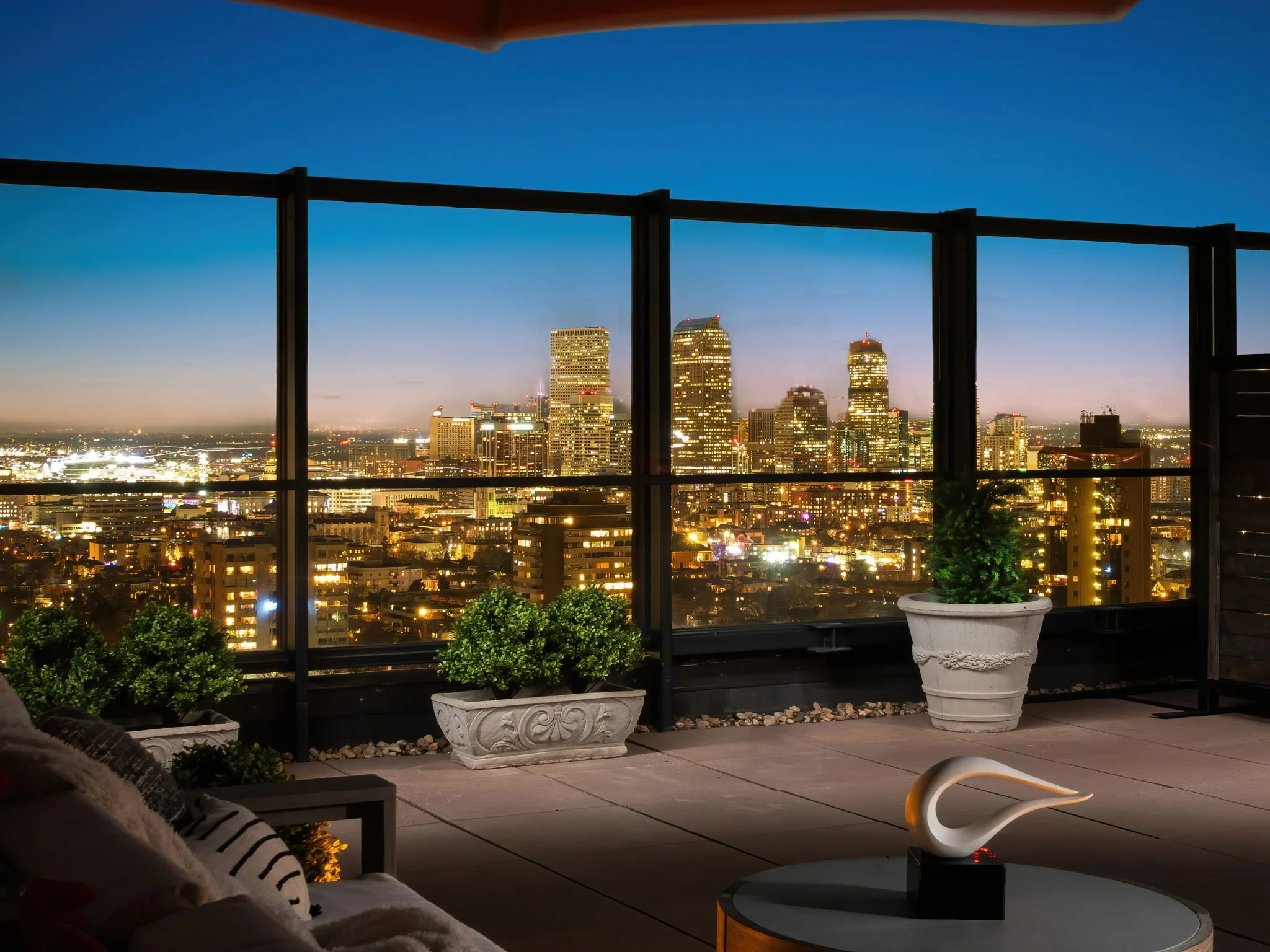 Nighttime city skyline view from a balcony, with tall illuminated skyscrapers in the background and potted plants on the balcony."