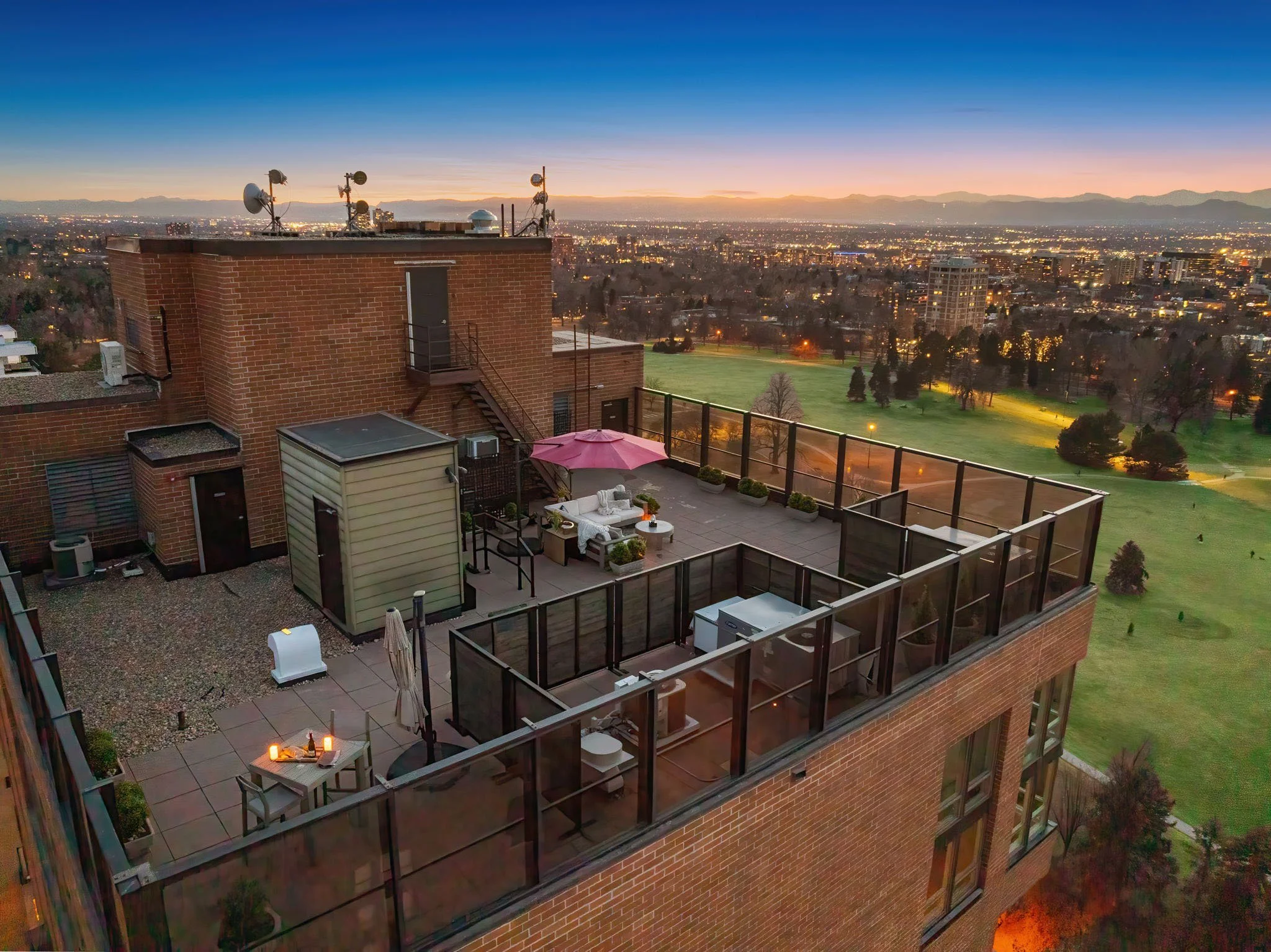 Rooftop terrace at sunset with outdoor seating, umbrella, and cityscape view in the background.