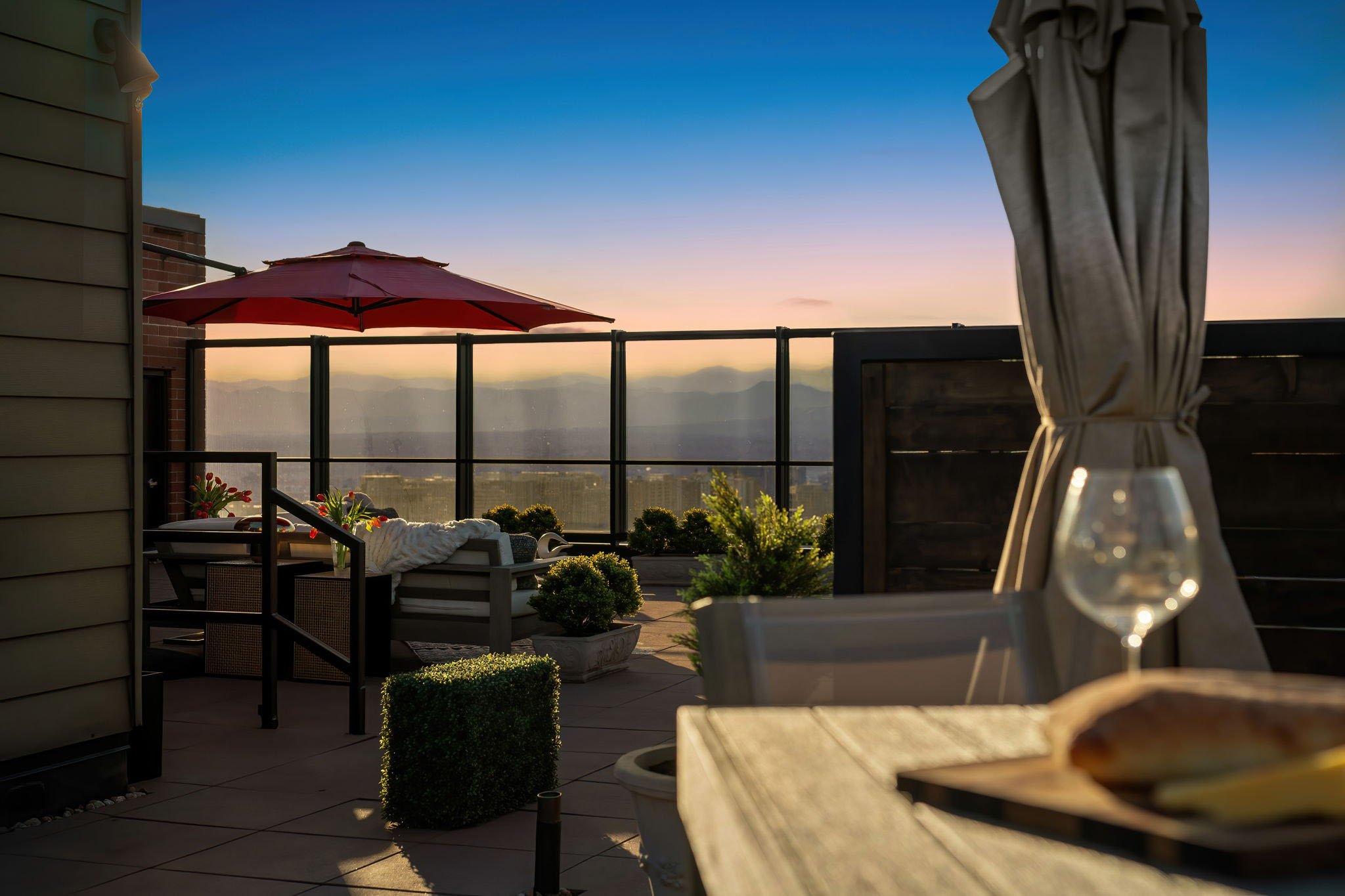 A rooftop patio during sunset with outdoor furniture, a red umbrella, potted plants, and a glass of wine on a table.