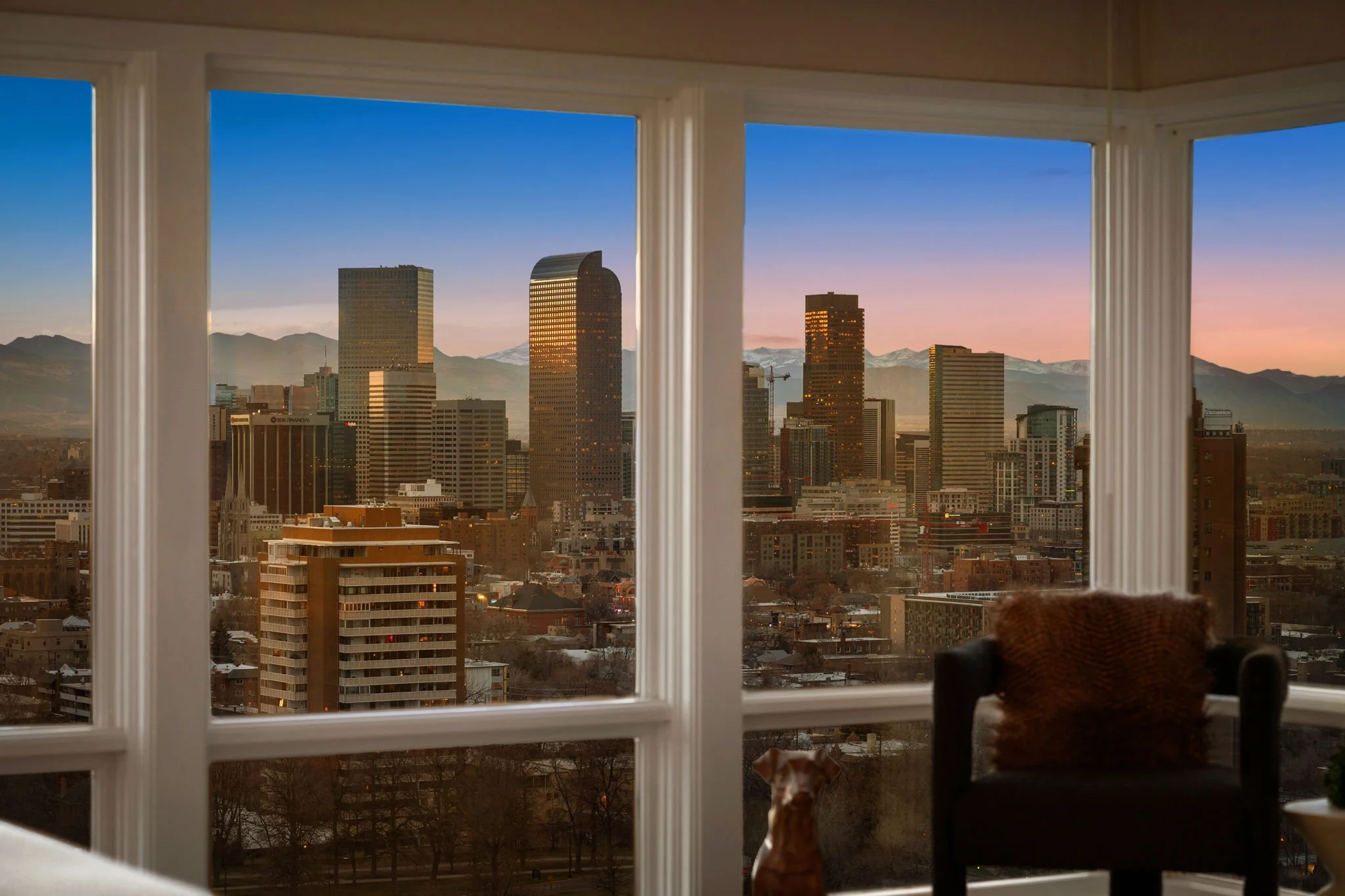 City skyline viewed through large window with mountains in the background during sunset, with a black chair and fur throw inside.