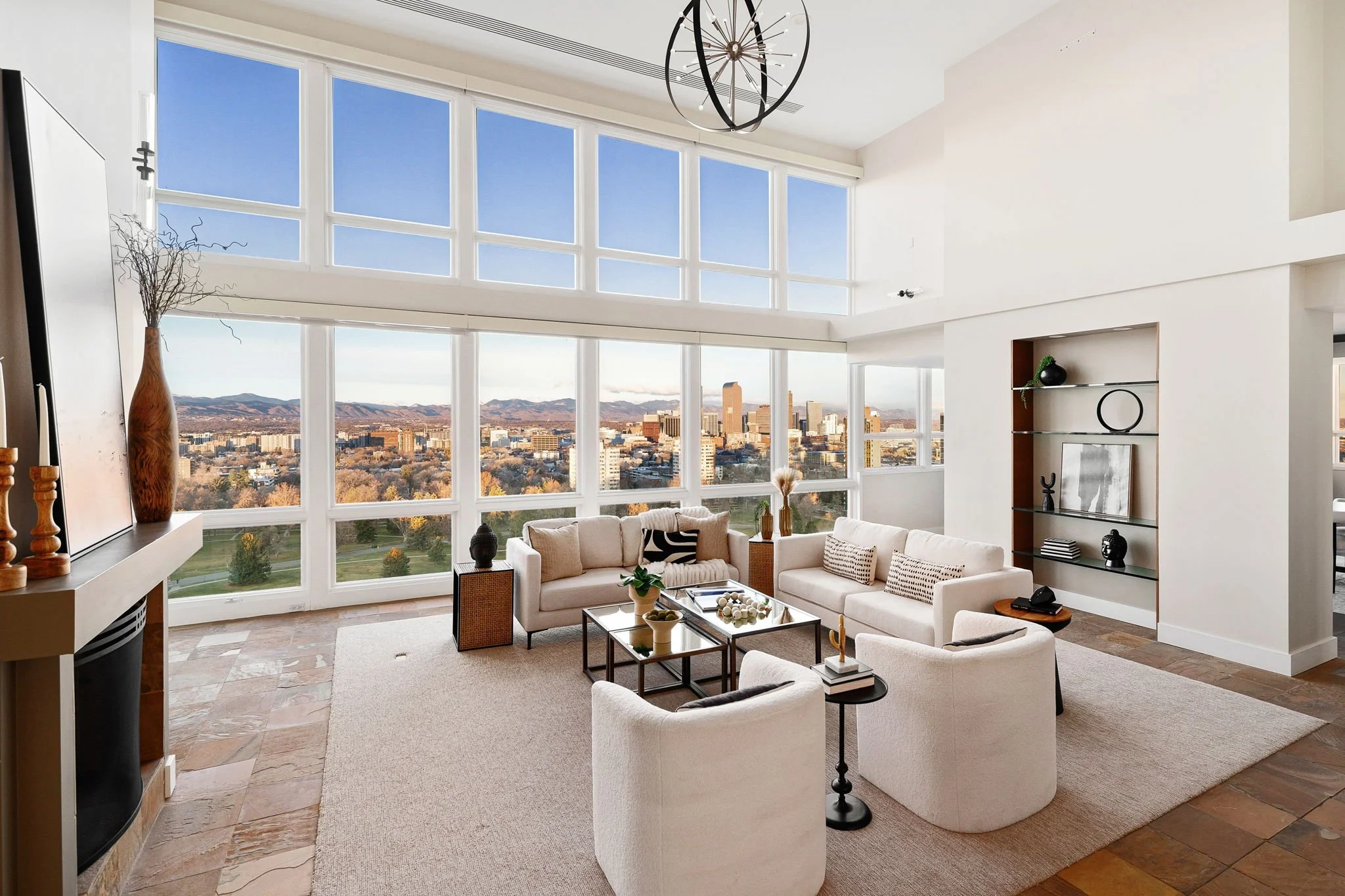 Living room with large windows showing a city view, white sofas, a glass coffee table with decorative items, and a built-in shelf with black and white decor.