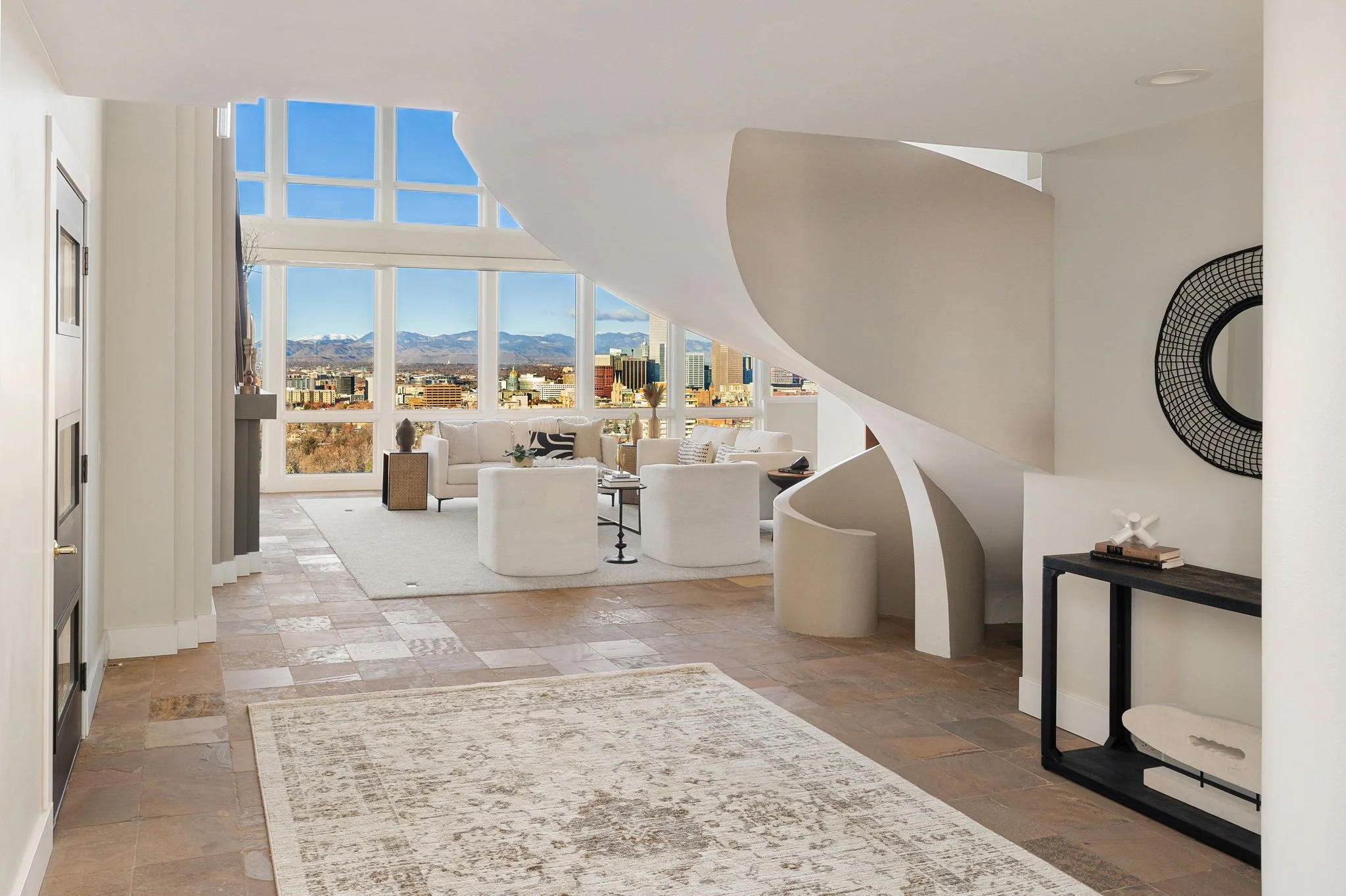 Modern living room with large windows showing cityscape and mountains, white curved staircase, white sofa and chairs, and decorative items.