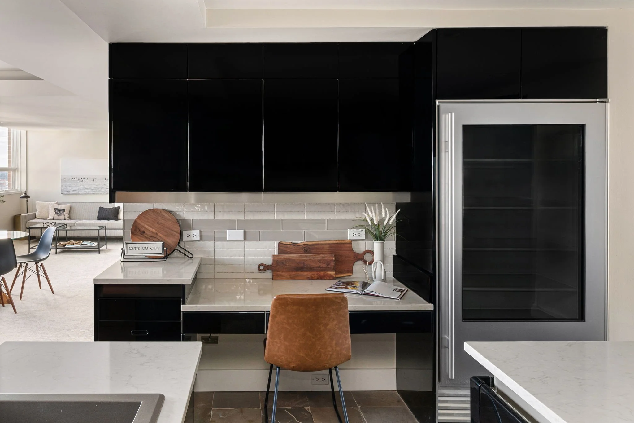 Modern kitchen with black cabinets, a stainless steel refrigerator, white marble countertops, and beige tiled backsplash. A brown chair is in front of the counter, and decorative items include a vase with white flowers, cutting boards, and an open ma