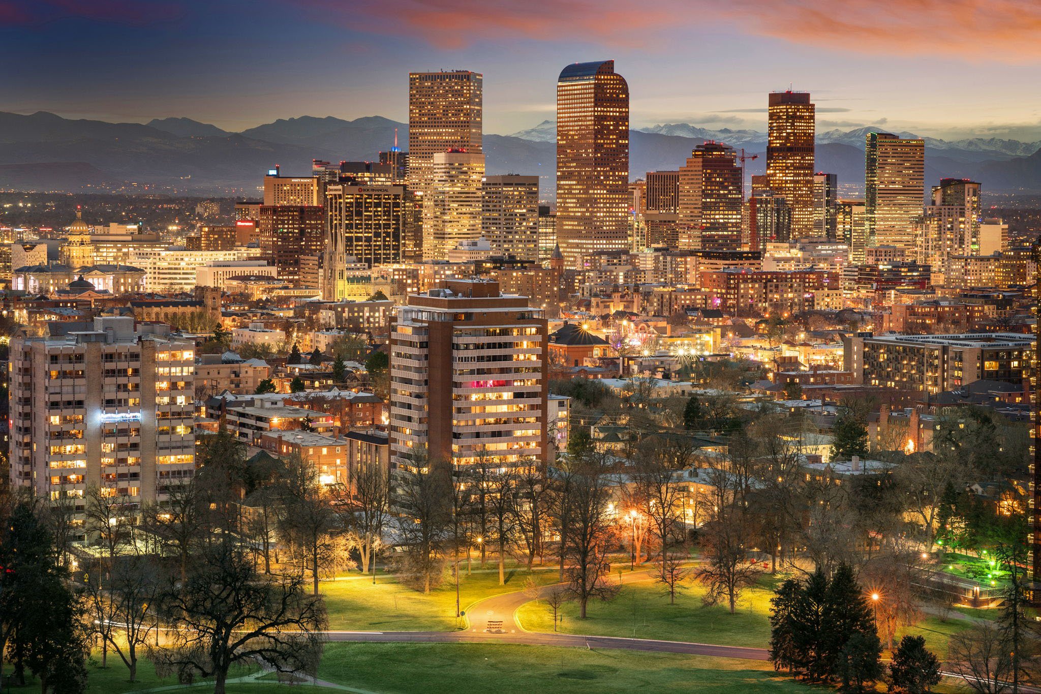 A city skyline at dusk with tall illuminated buildings, mountains in the background, and a park with trees and walking paths in the foreground.