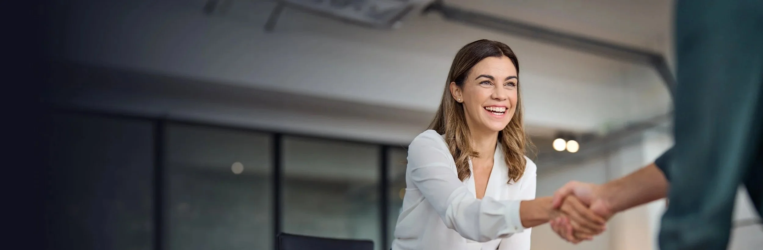 A woman smiling and shaking hands with another person in a professional office setting.