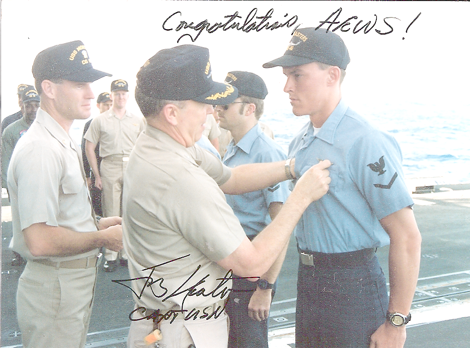 An older man in a beige uniform pins a young man in a blue military uniform with a black cap on the chest while on a ship, with a line of soldiers in uniform behind them. The scene is outdoors near the ocean, with handwritten text saying 'Congratulations HCWS!' and a signature.