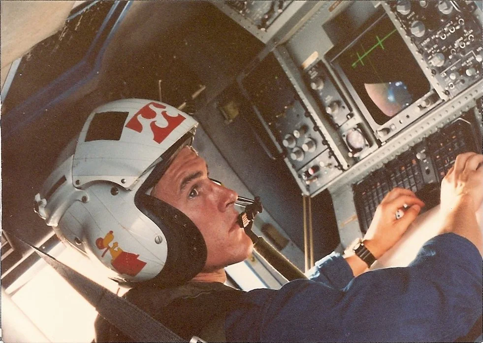 A man wearing a vintage racing helmet with the number 33 on it is sitting in the cockpit of an aircraft or spacecraft, surrounded by control panels and instruments.
