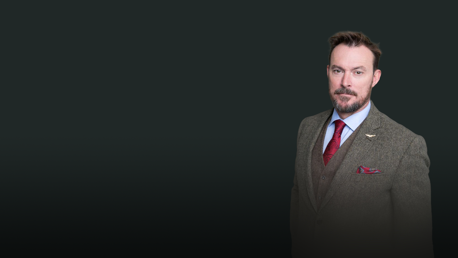 A man with brown hair, beard, and mustache wearing a brown patterned suit, light blue shirt, red tie with paisley pattern, and a pocket square. He has a small pin on his lapel. The background is dark and plain.